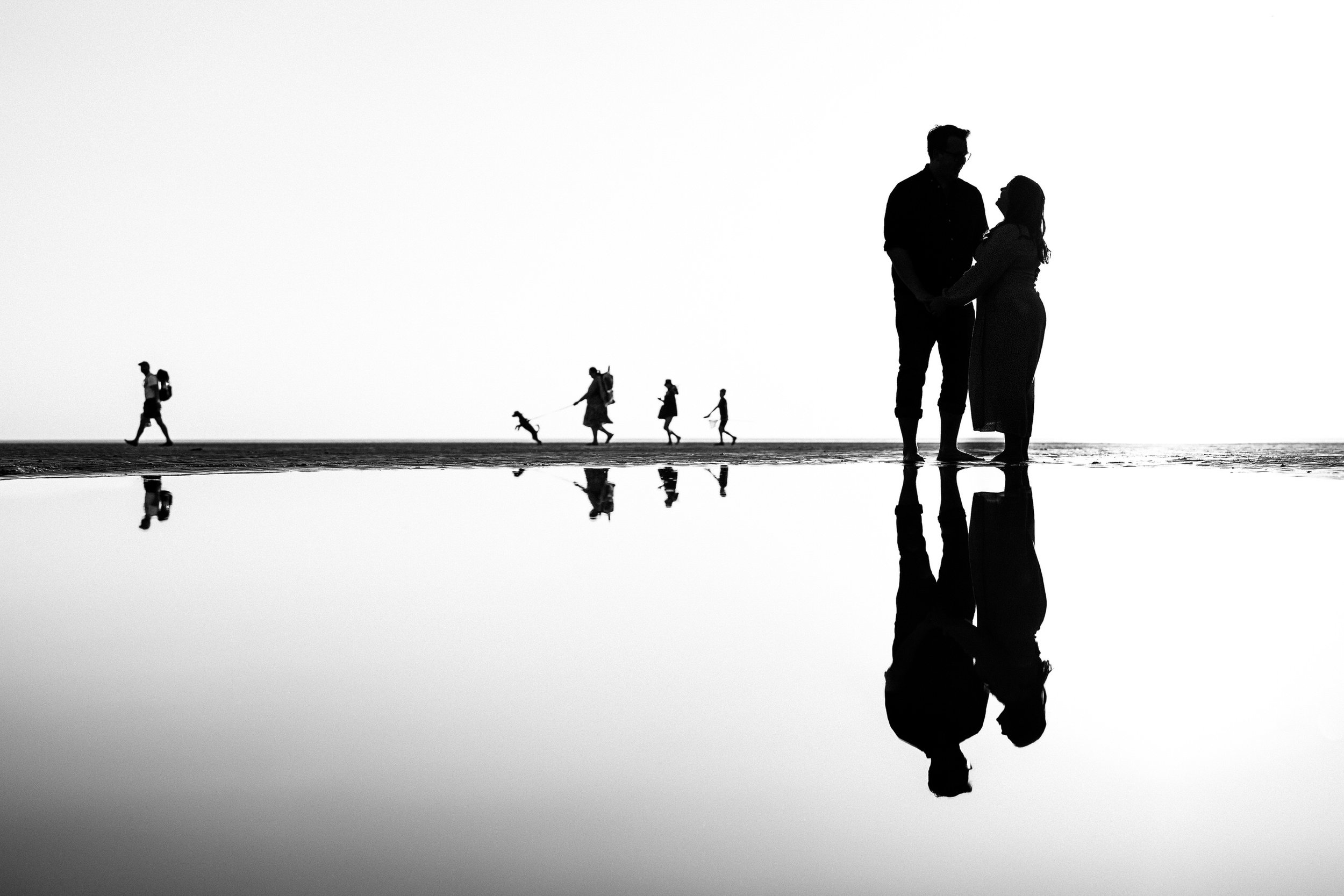 Black-and-white beach silhouette of a couple standing in shallow water, with their reflections and distant beachgoers.