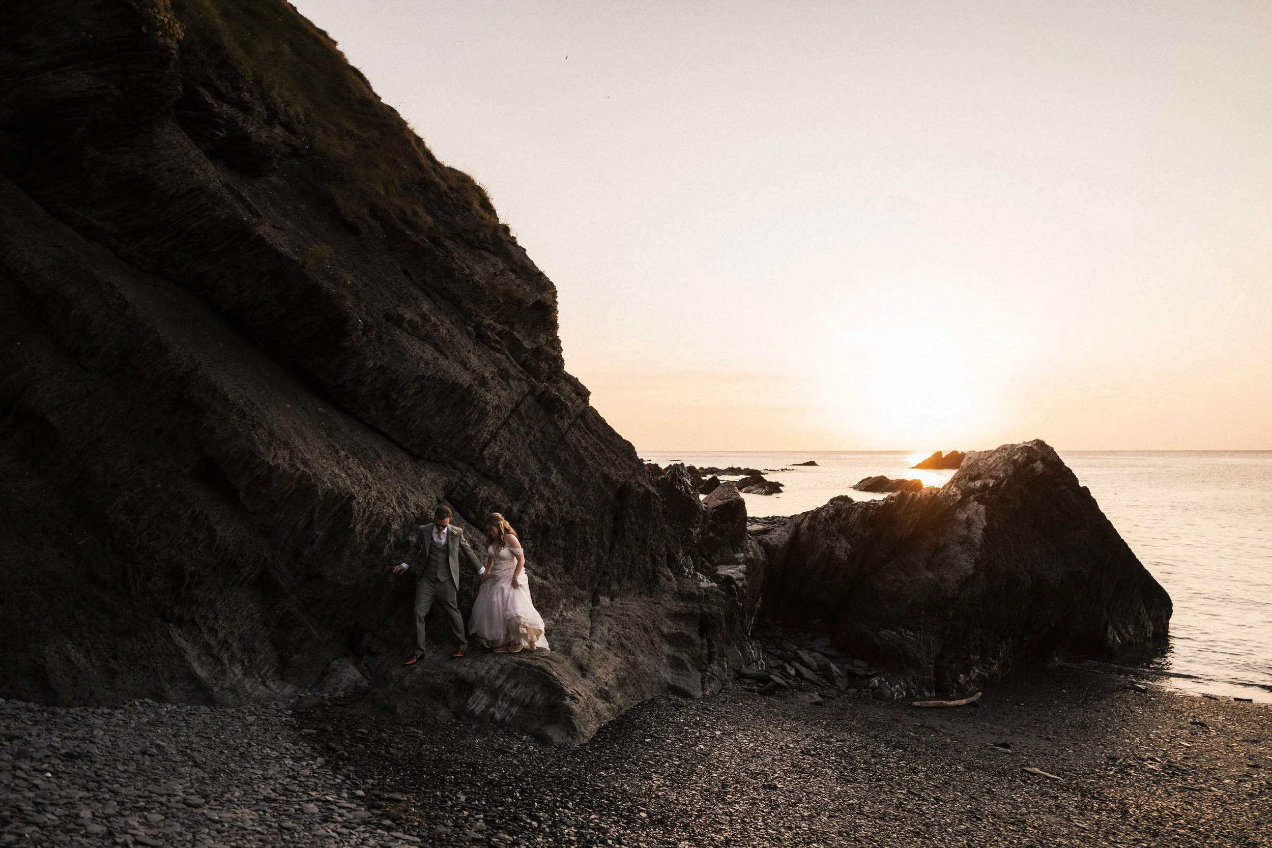 Bride and groom walking among dark coastal cliffs at sunset