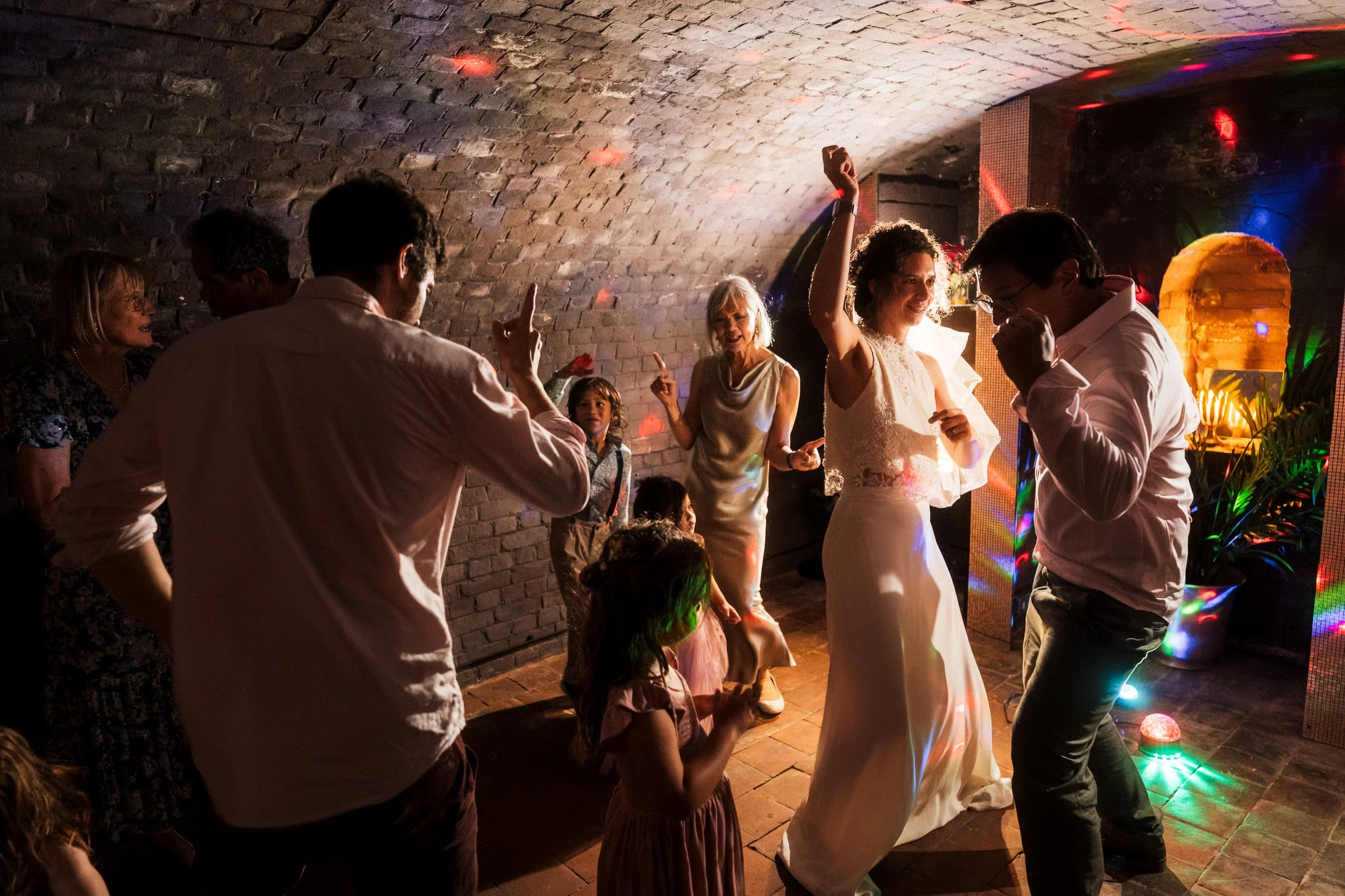 Bride and wedding guests dancing under colorful party lights in a brick-walled reception venue