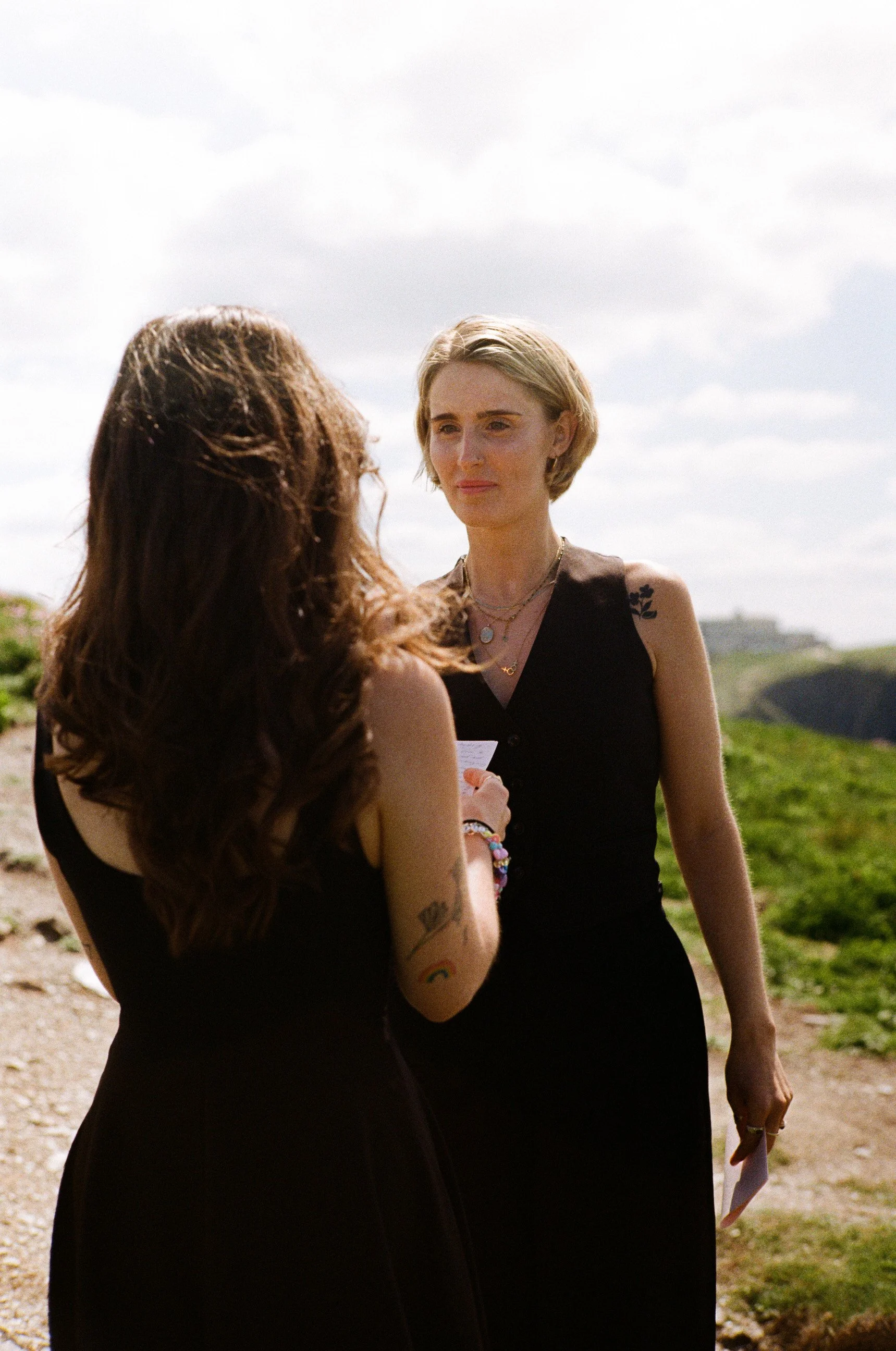 Two women in black dresses talking outdoors against a bright sky and green landscape