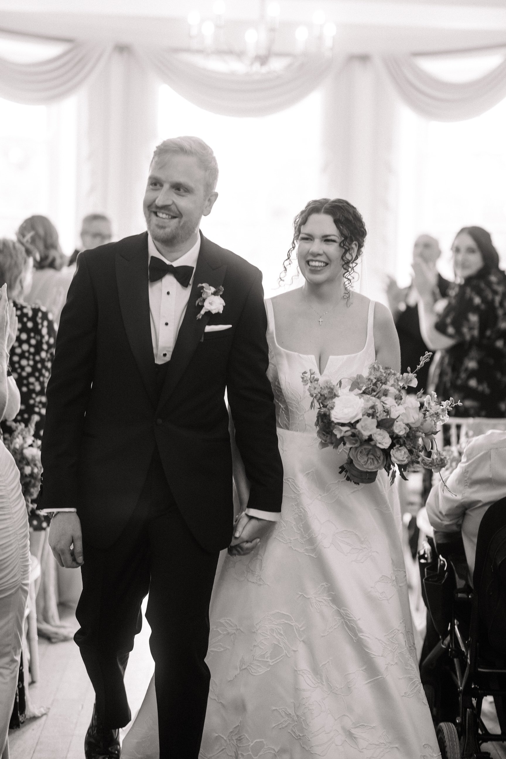 Black-and-white wedding photo of a smiling bride and groom walking down the aisle, surrounded by guests.