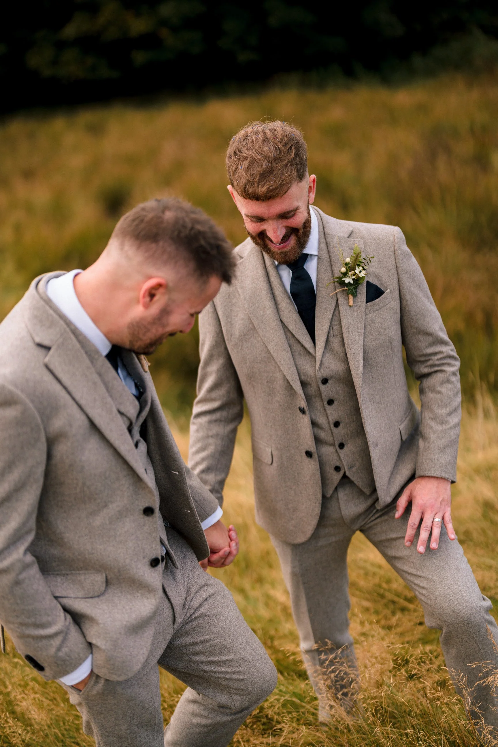 Two men in gray suits laughing outdoors, one wearing a floral boutonniere.