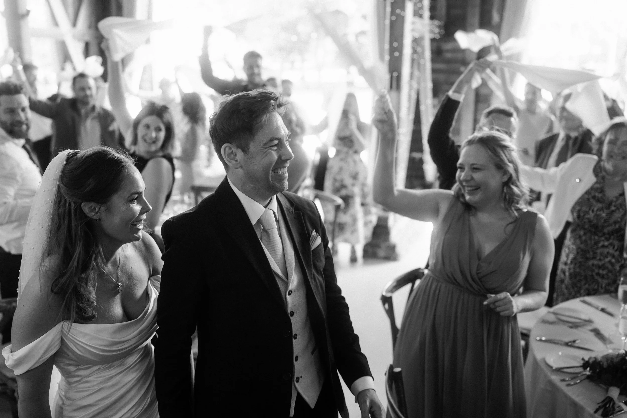 Bride and groom smiling at a lively wedding reception surrounded by cheering guests