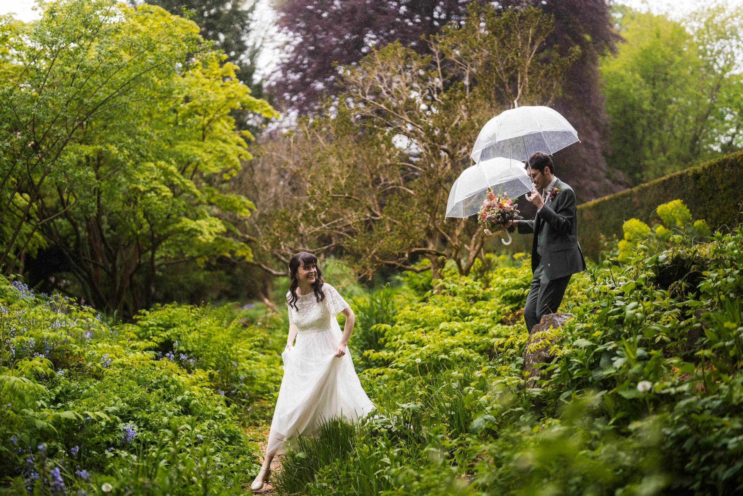Bride and groom walking through a lush garden in the rain under clear umbrellas