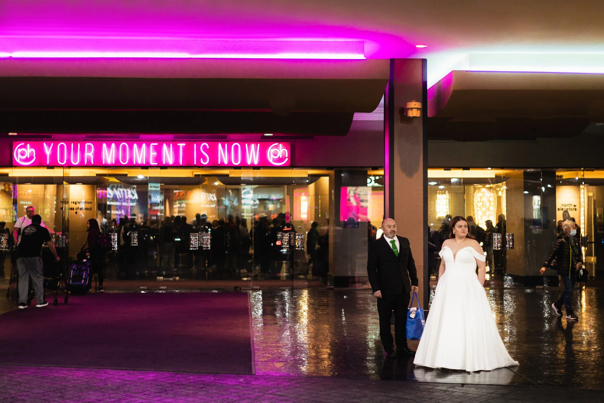 Bride and groom outside a theater entrance beneath a neon “Your Moment Is Now” sign