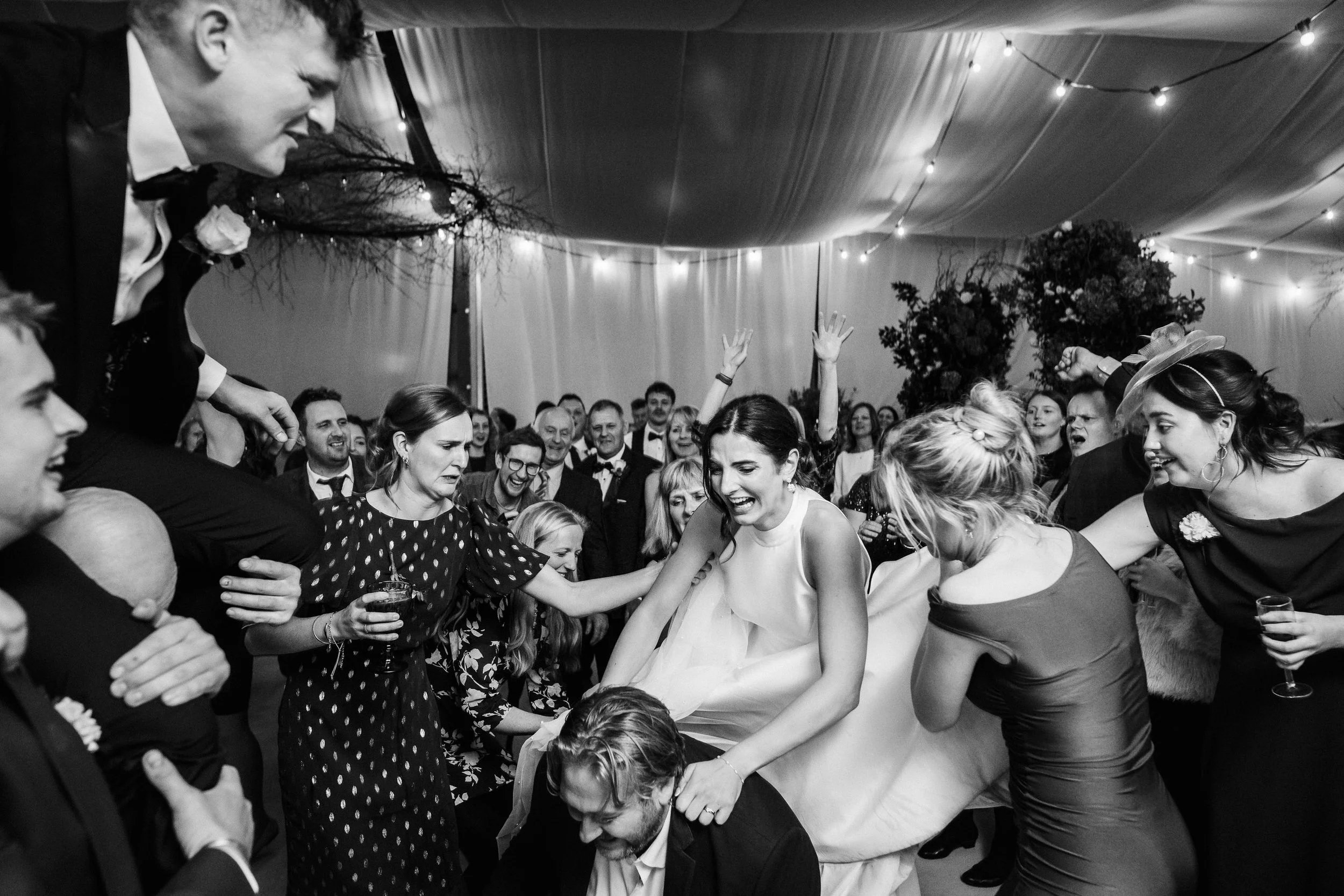 Black-and-white wedding reception photo of a laughing bride surrounded by dancing guests beneath string lights.