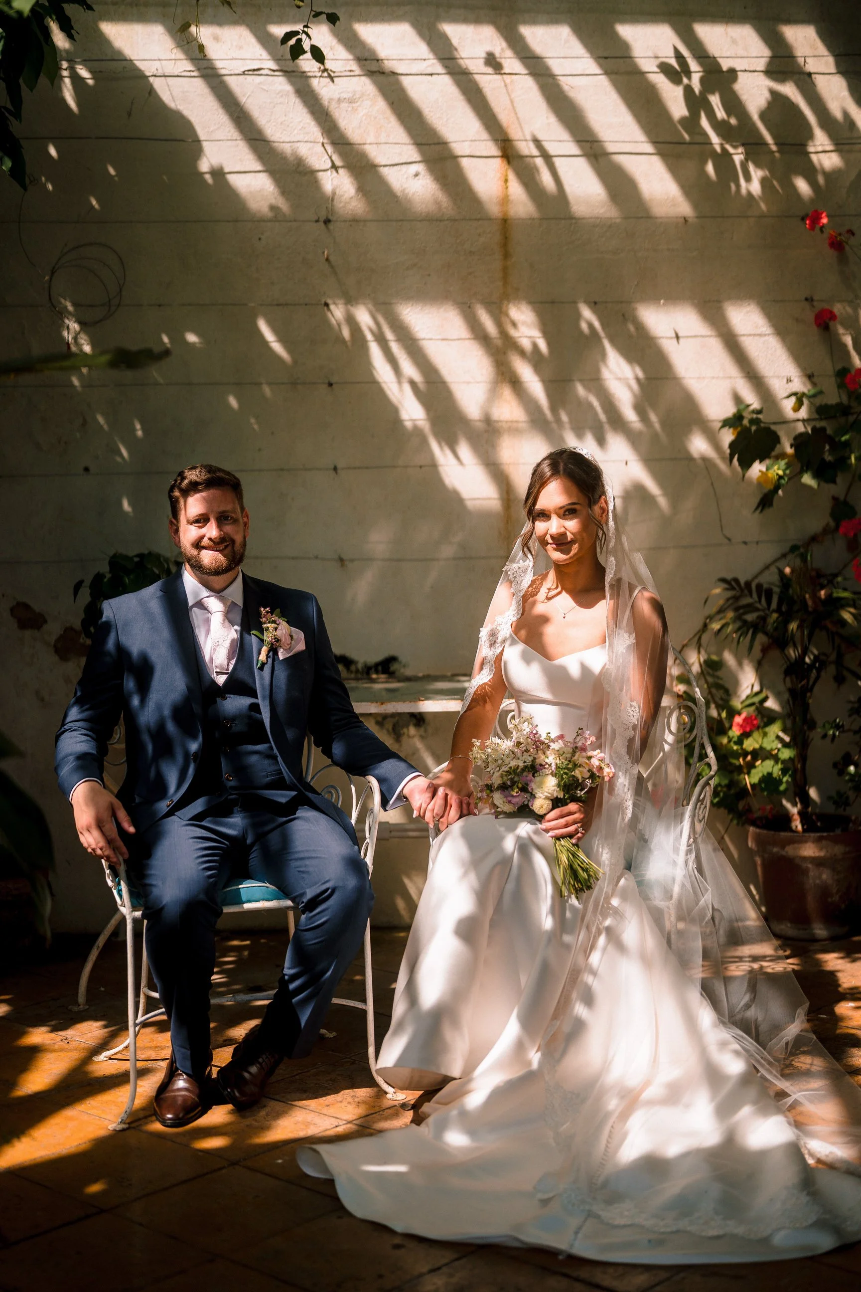 Newlywed couple seated together, with the groom in a navy suit and the bride in a white wedding dress and veil holding a bouquet.