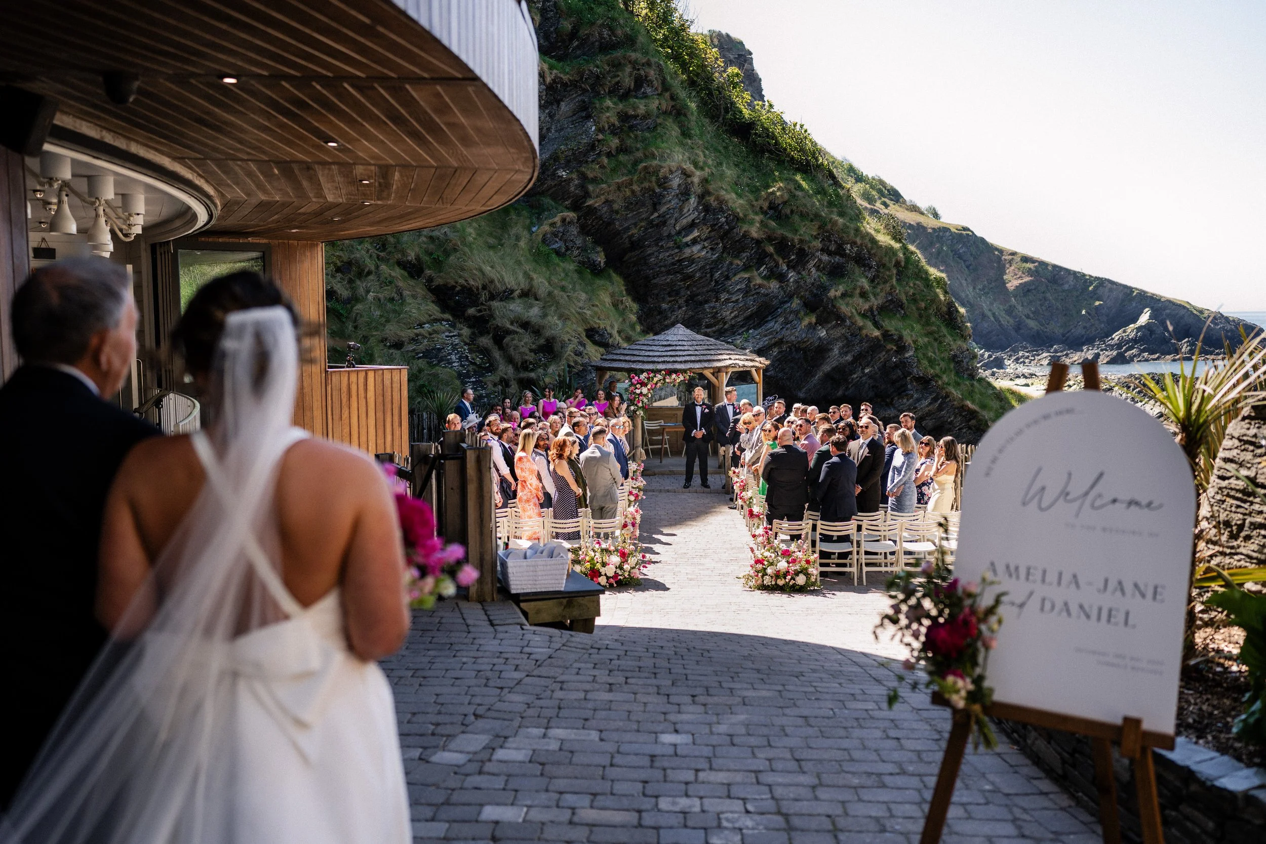 Bride walking toward an outdoor wedding ceremony overlooking a rugged mountain and seaside landscape