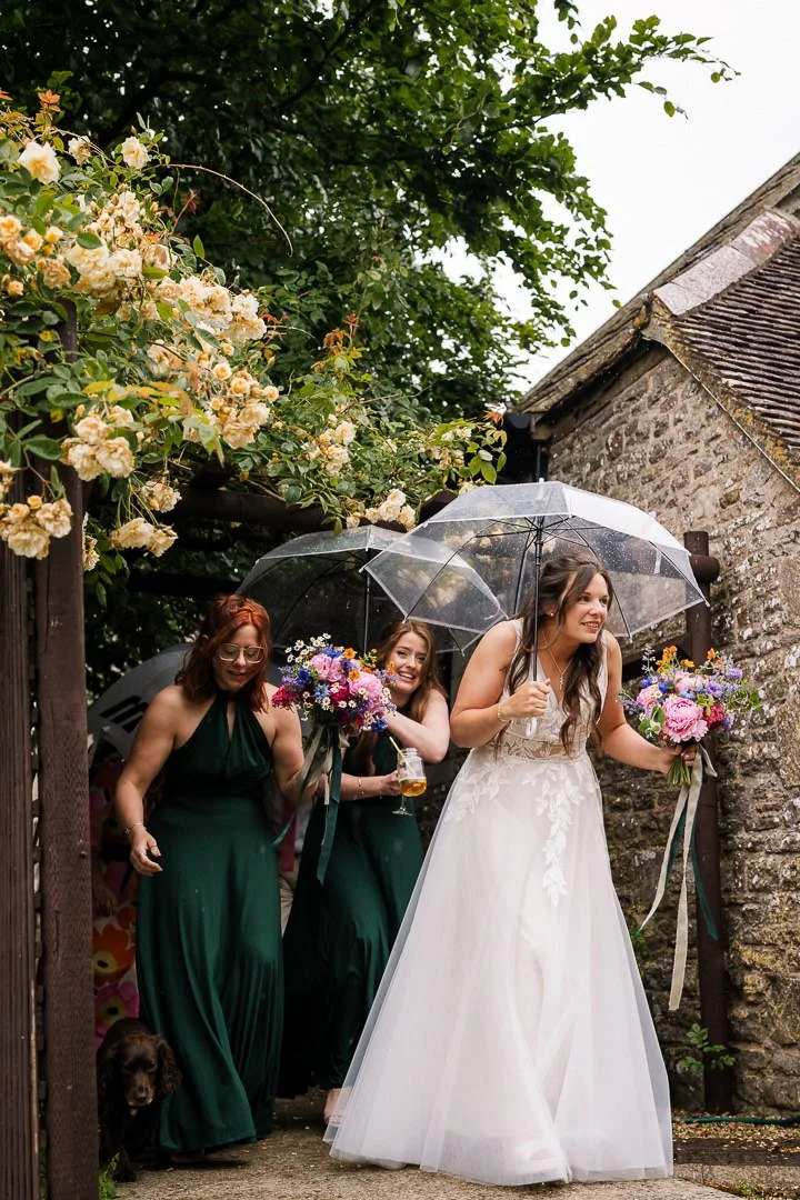 Bride and bridesmaids holding bouquets and clear umbrellas outside a rustic stone building in the rain