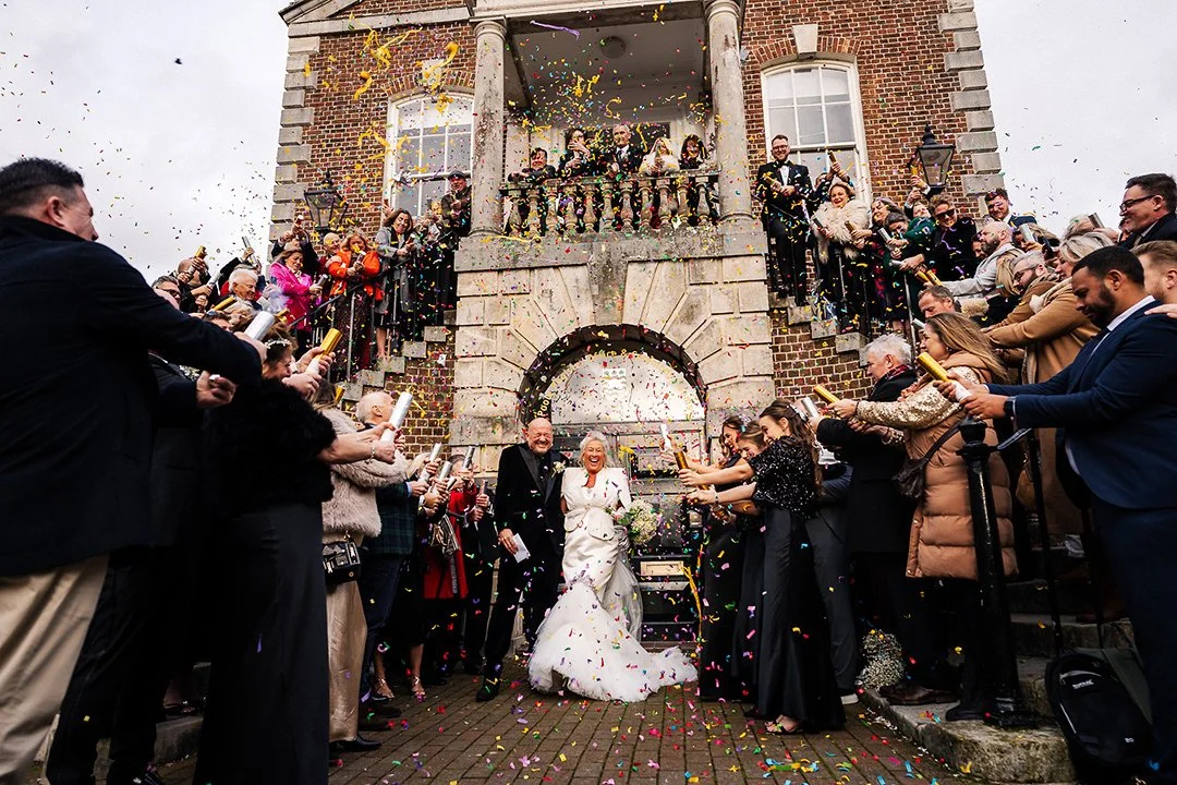 Newlywed couple exits a historic brick building as guests shower them with colorful confetti
