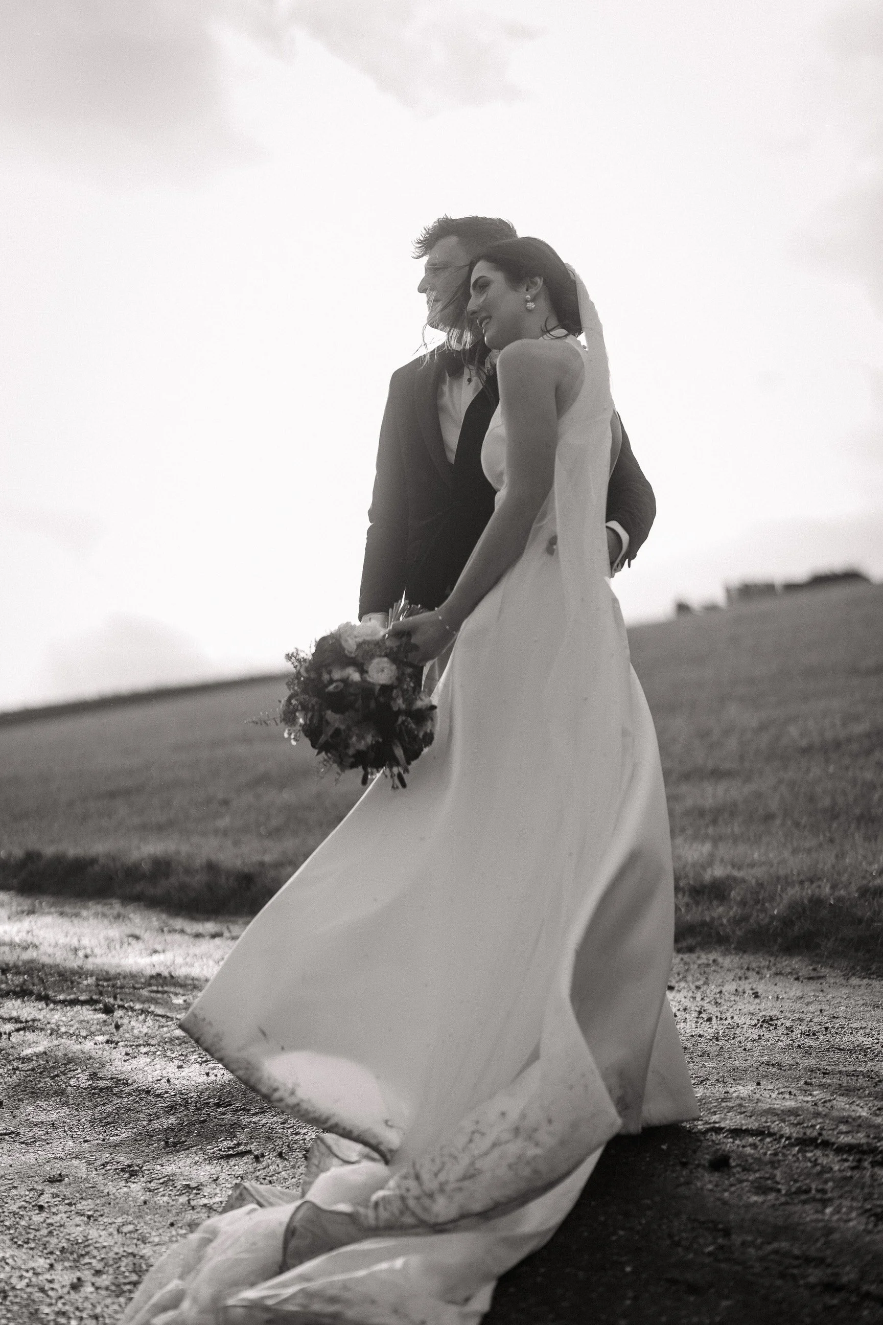 Black-and-white outdoor wedding portrait of a bride in a flowing gown holding a bouquet with her groom behind her.
