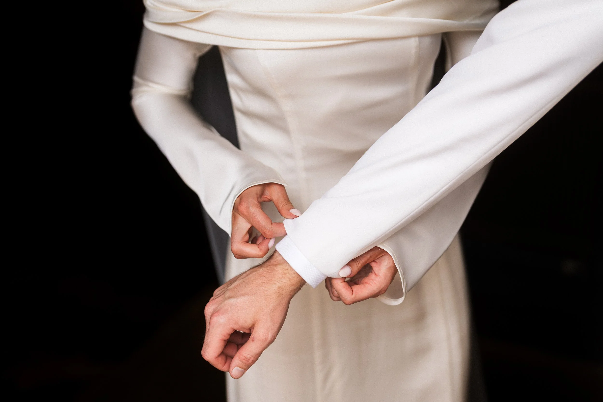 Close-up of a person in a white suit adjusting their cufflink against a black background
