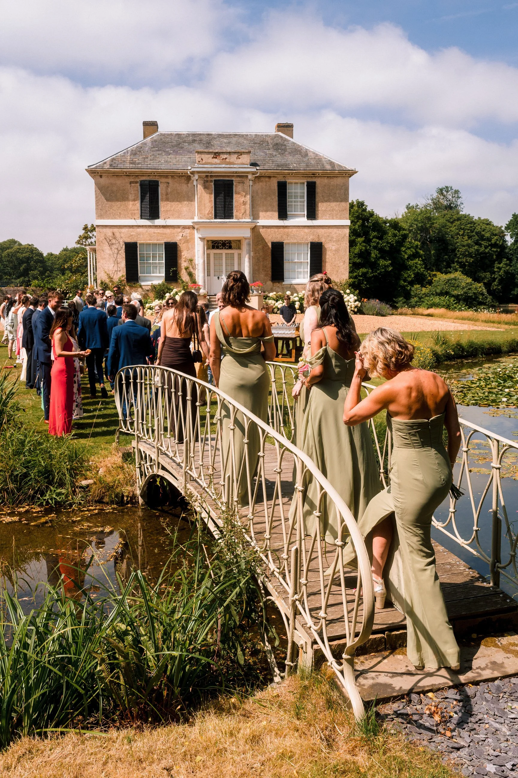 Wedding guests gather outside a historic country house beside a lily pond and ornamental footbridge.