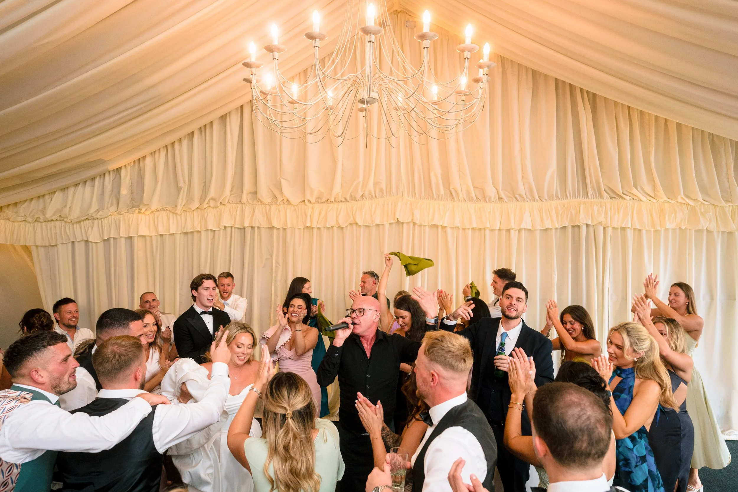 Wedding guests celebrating beneath a large candlelit chandelier in a draped reception tent