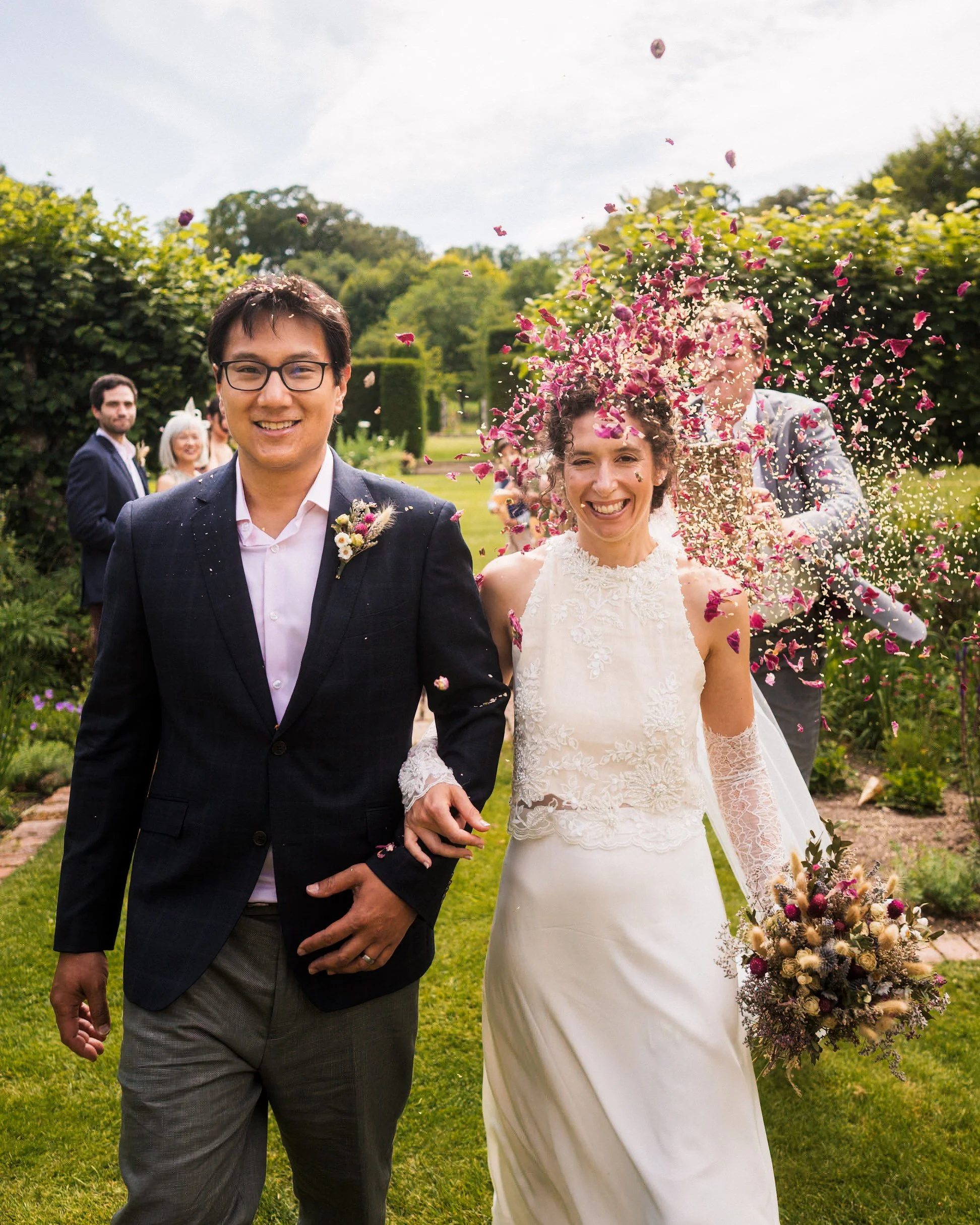 Newlywed couple smiling in a garden as wedding guests throw pink flower petals