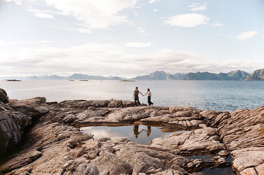 Couple holding hands on rocky shoreline beside a tide pool, with calm sea and mountain range in the background