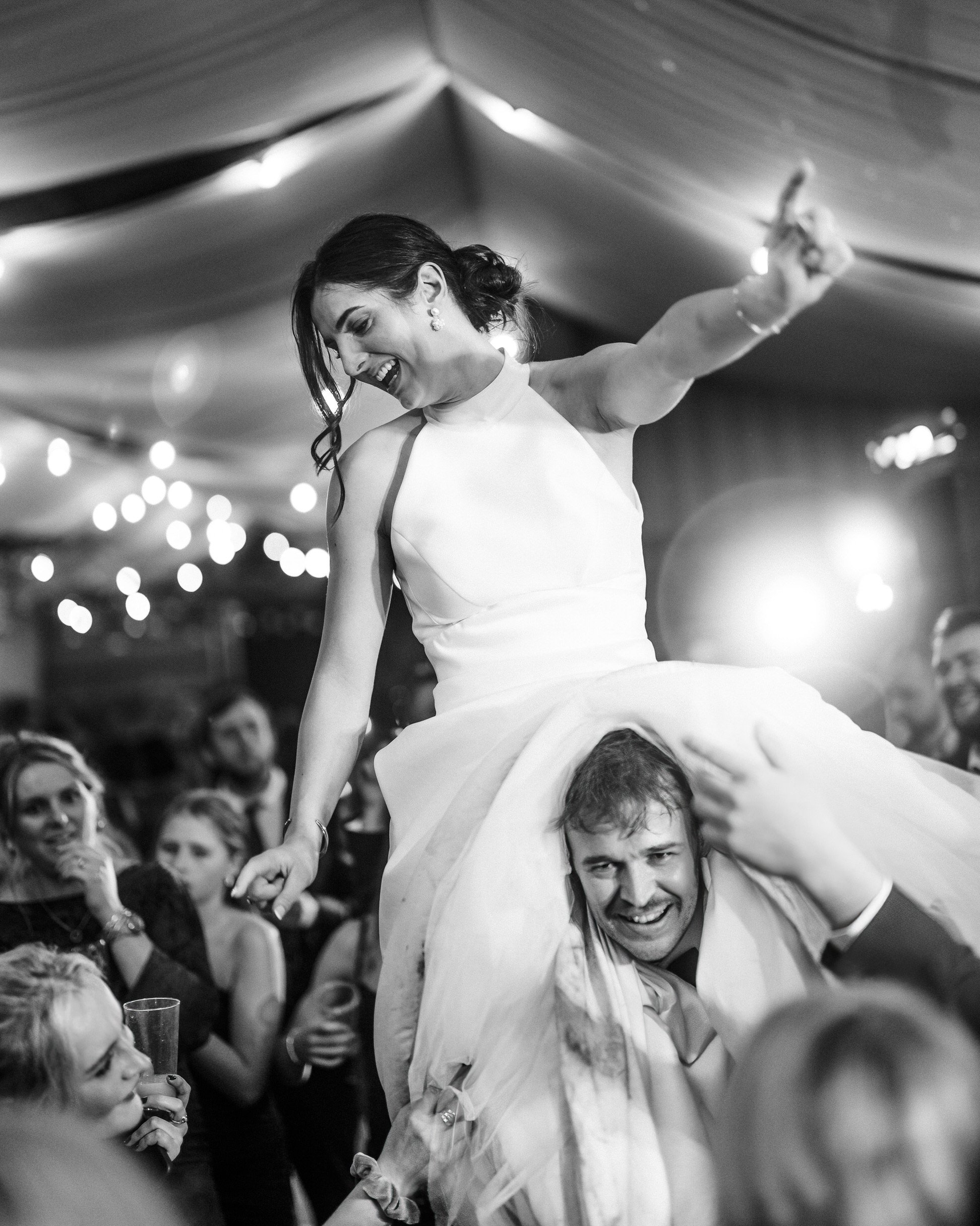 Black-and-white wedding reception photo of a smiling bride riding on a groom’s shoulders as guests celebrate around them.