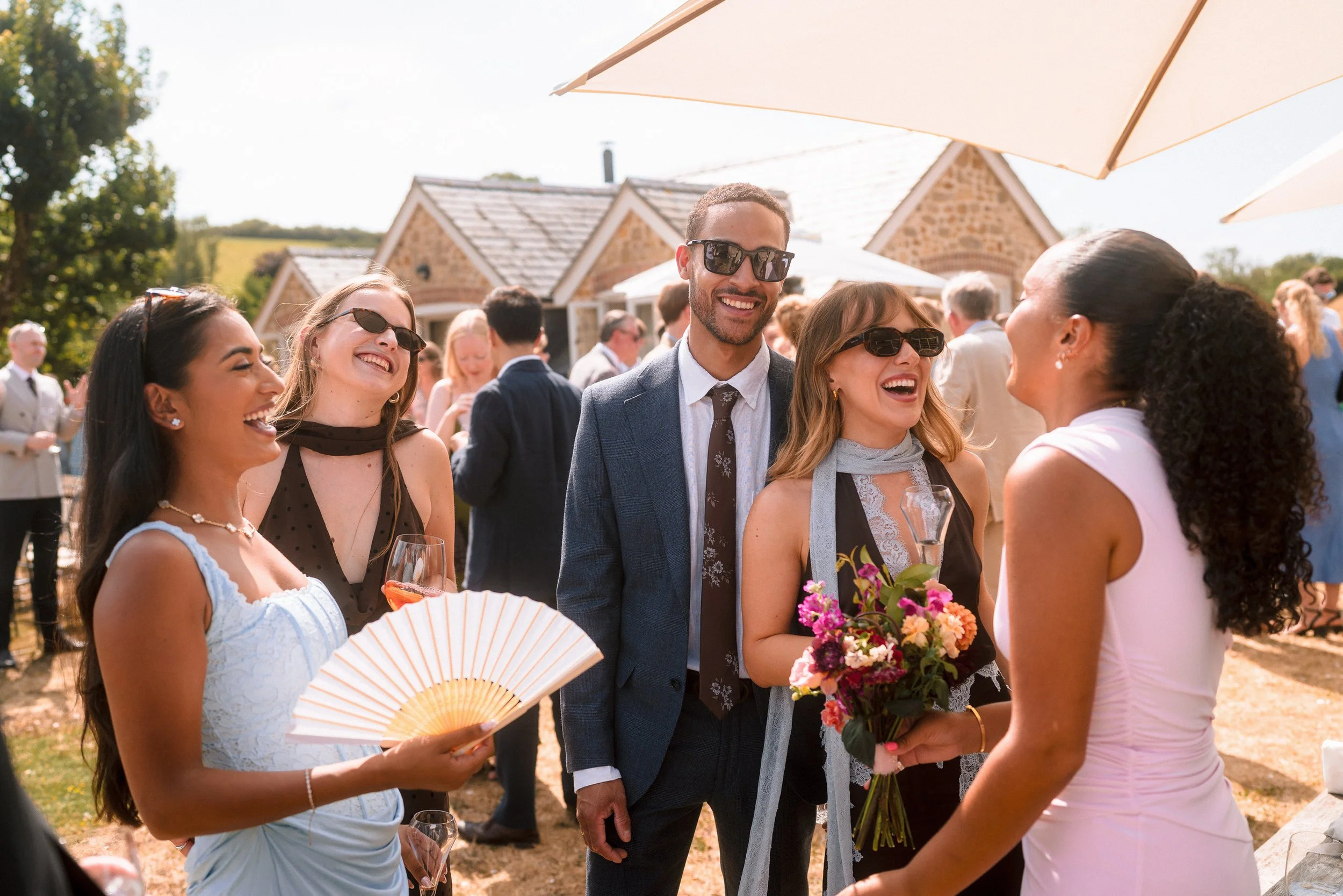 Friends laughing together at a sunny outdoor wedding reception with drinks and flowers