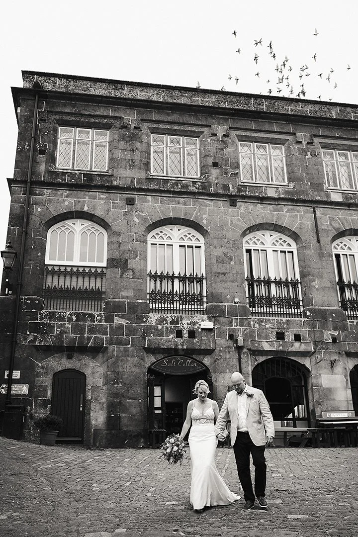 Black-and-white wedding photo of a bride and groom walking on a cobblestone street in front of a historic stone building, with birds flying overhead