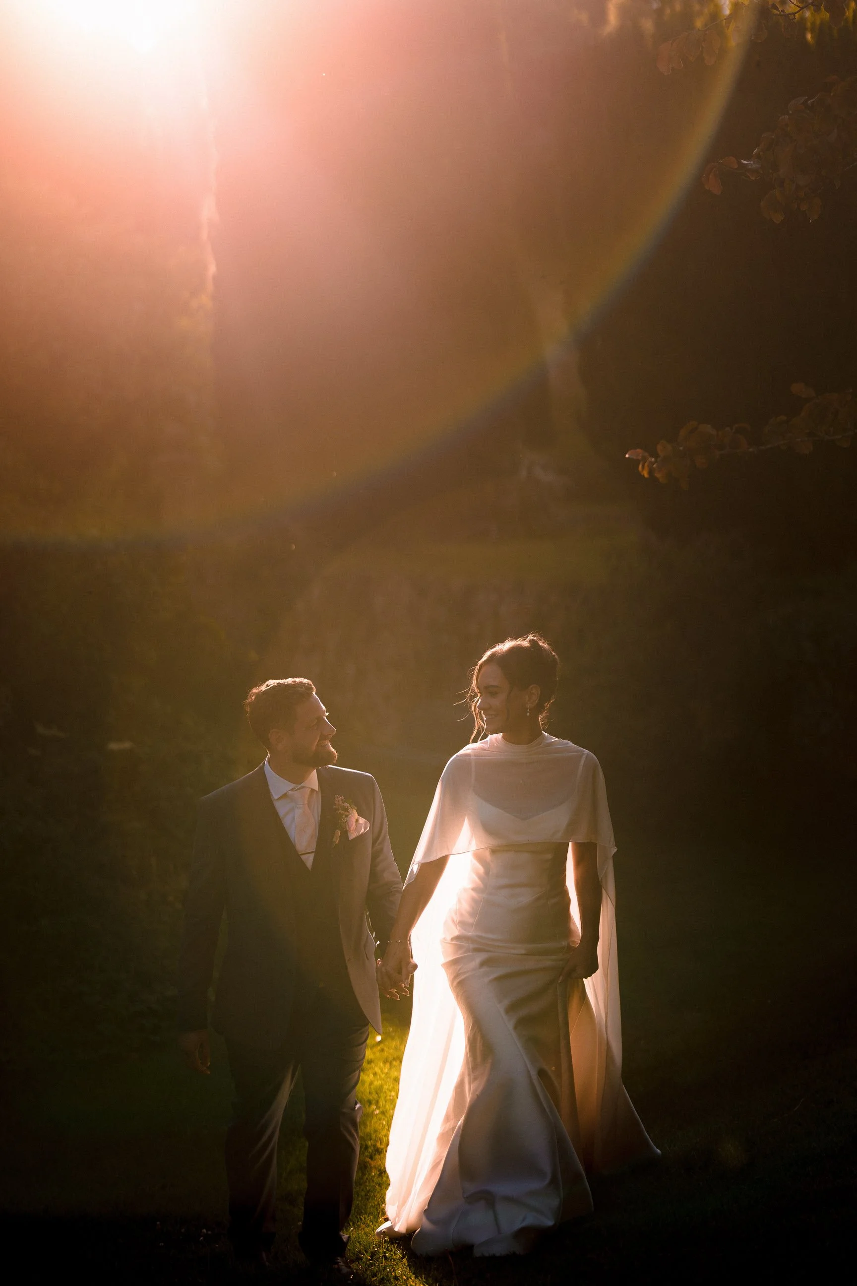 Newlywed couple holding hands and walking through a sunlit garden, the groom in a dark suit and the bride in a flowing white wedding dress.