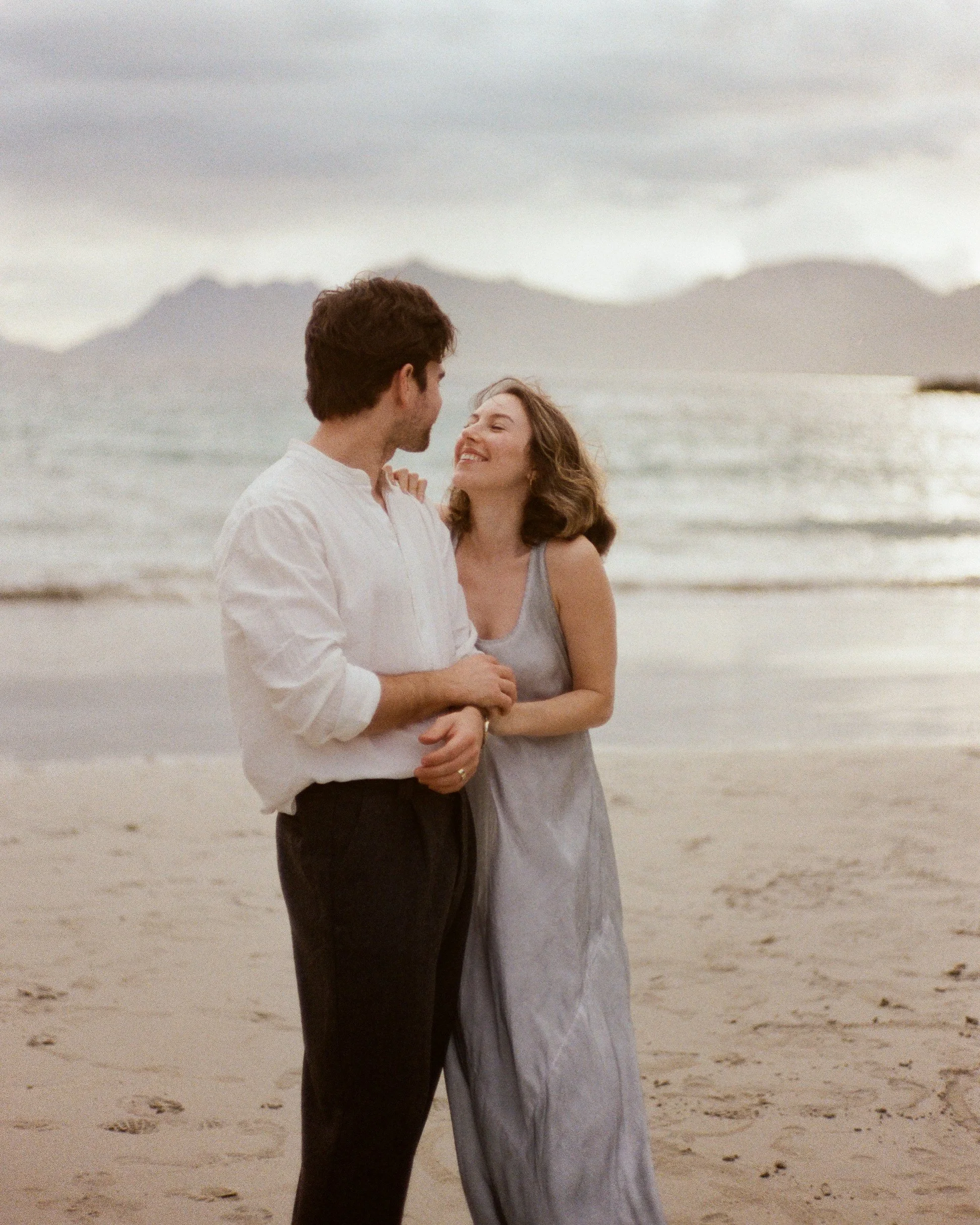 Smiling couple embracing on a sandy beach with ocean waves and mountains in the background