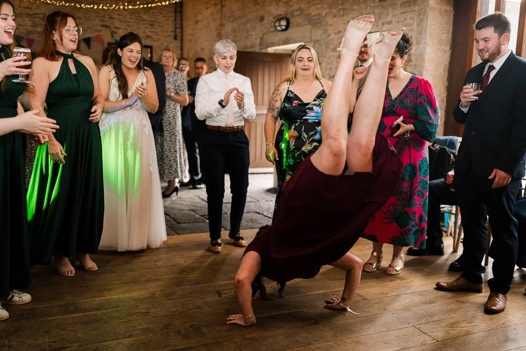 Wedding guests cheer as a person performs a playful handstand on the dance floor.