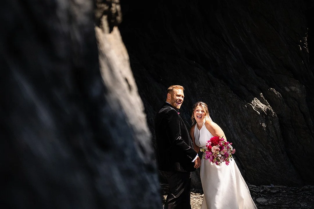 Newlywed couple smiling in a rocky cave, with the bride holding a colorful bouquet.