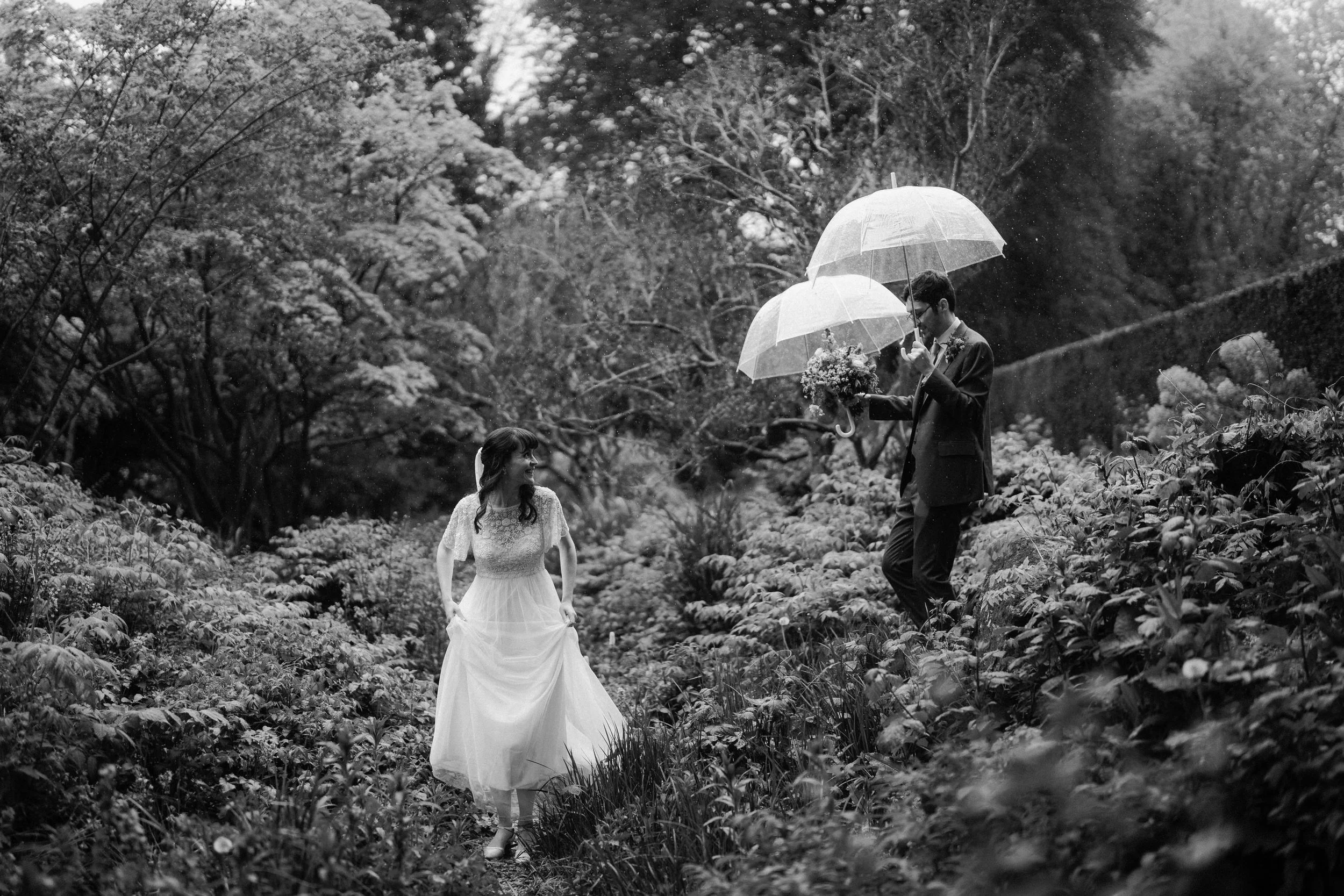 Black-and-white wedding photo of a bride and groom holding clear umbrellas in a rainy forest.