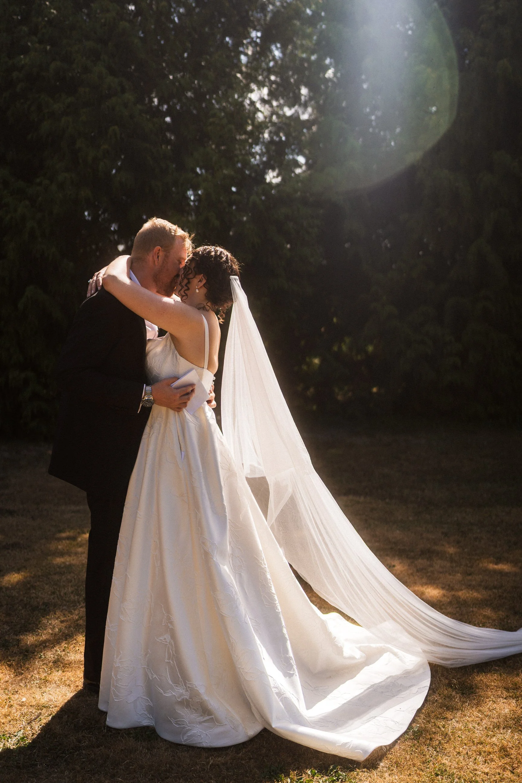 Newlywed couple embracing outdoors in warm sunlight, with the bride’s long wedding dress train spread across the grass