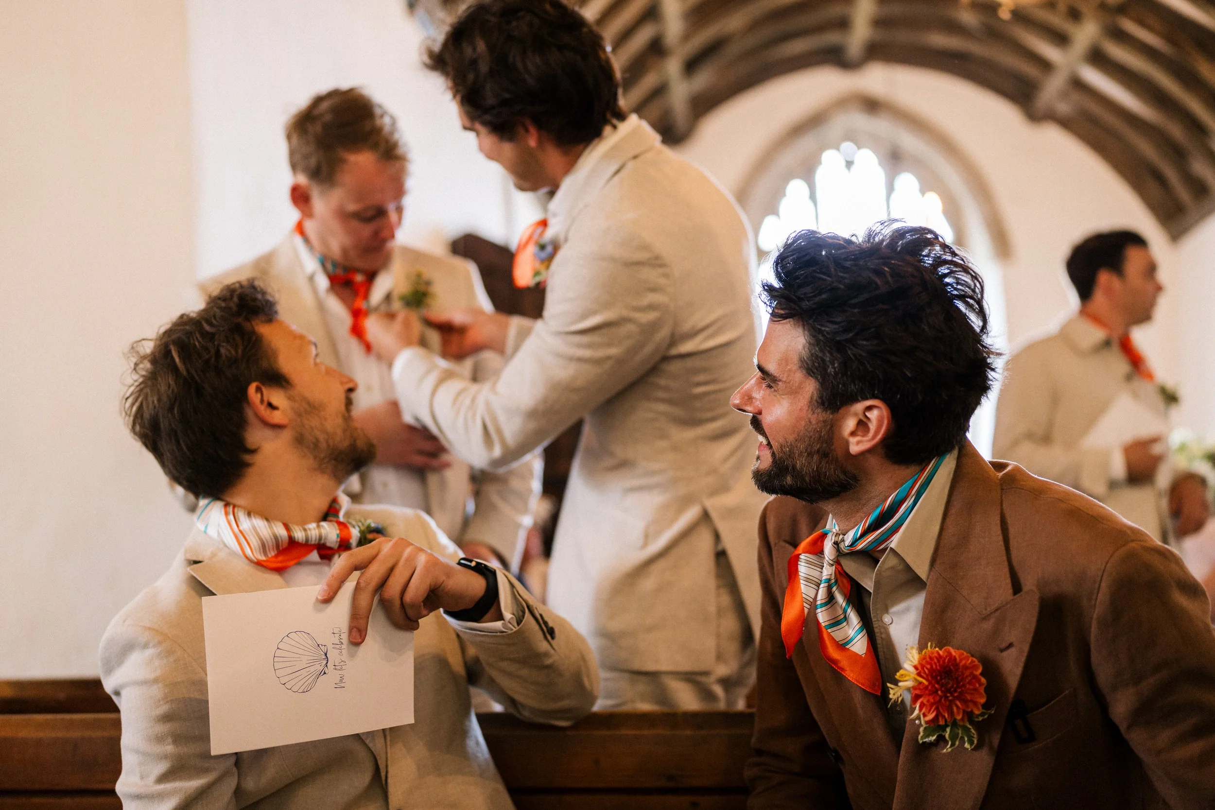 Wedding guests in colorful scarves and boutonnières gather inside a church, with one man holding a seashell-illustrated program.