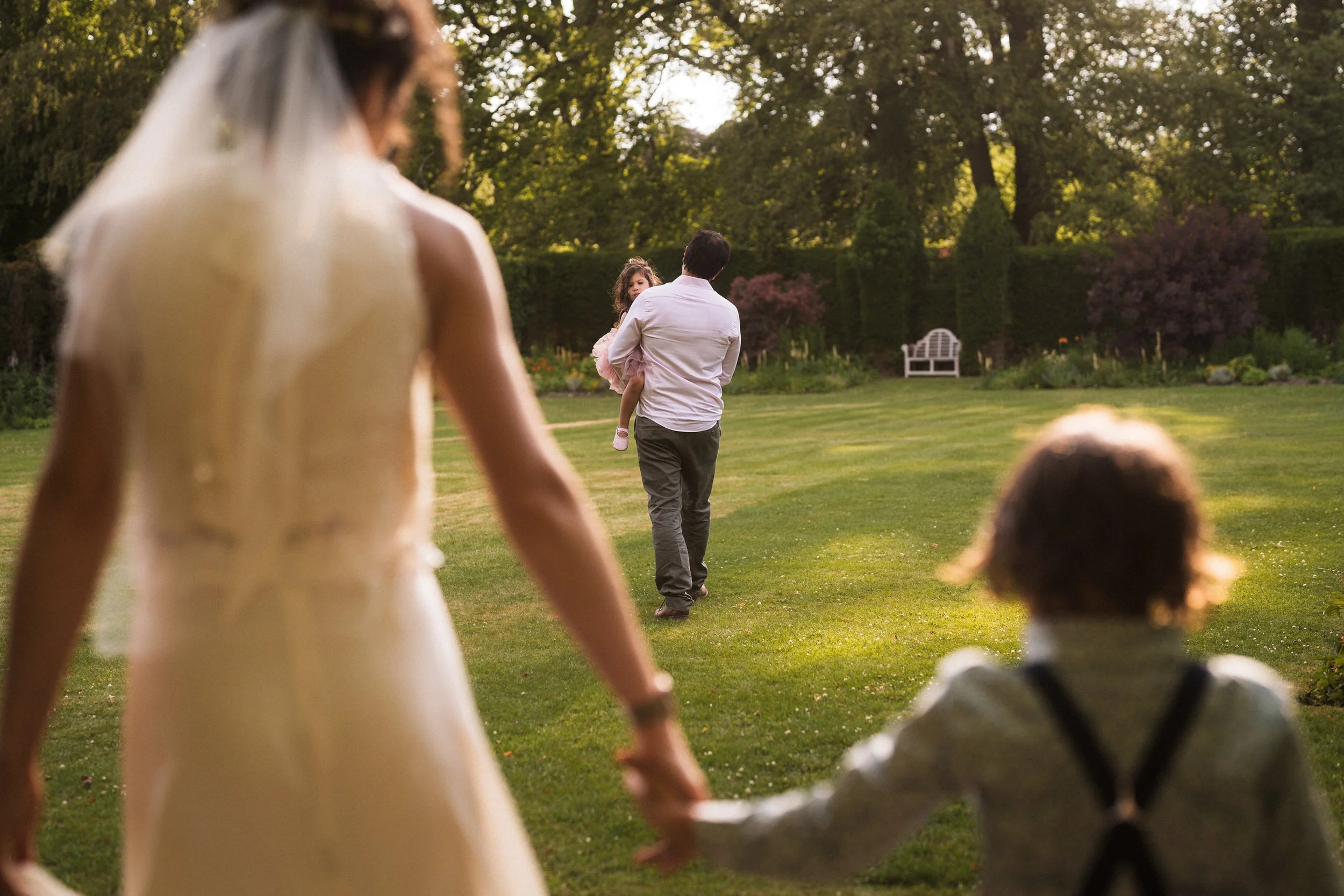 Bride and groom walking through a garden with a young child, viewed from behind.