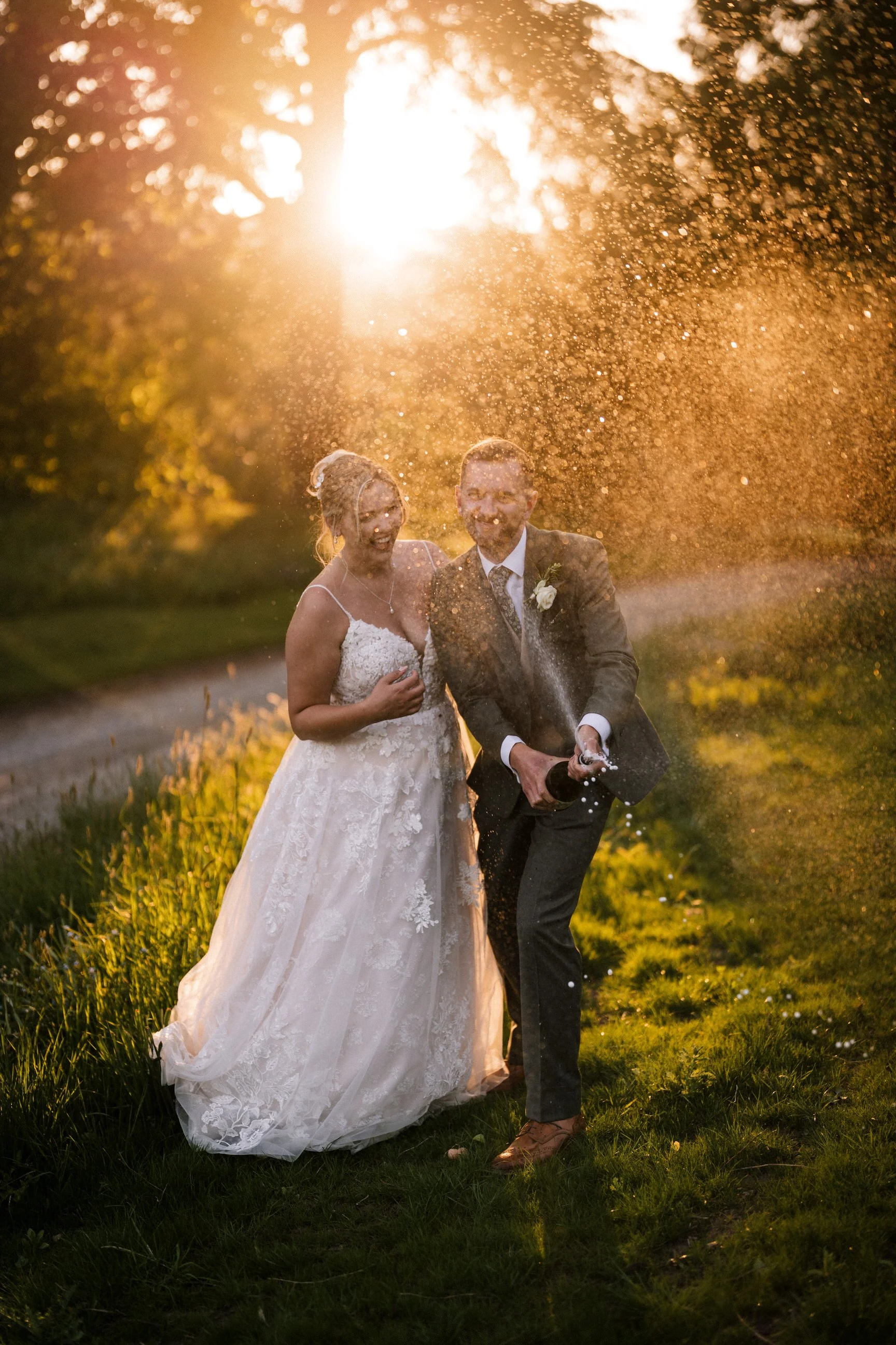 Newlywed couple smiling outdoors as champagne sprays around them in golden sunset light