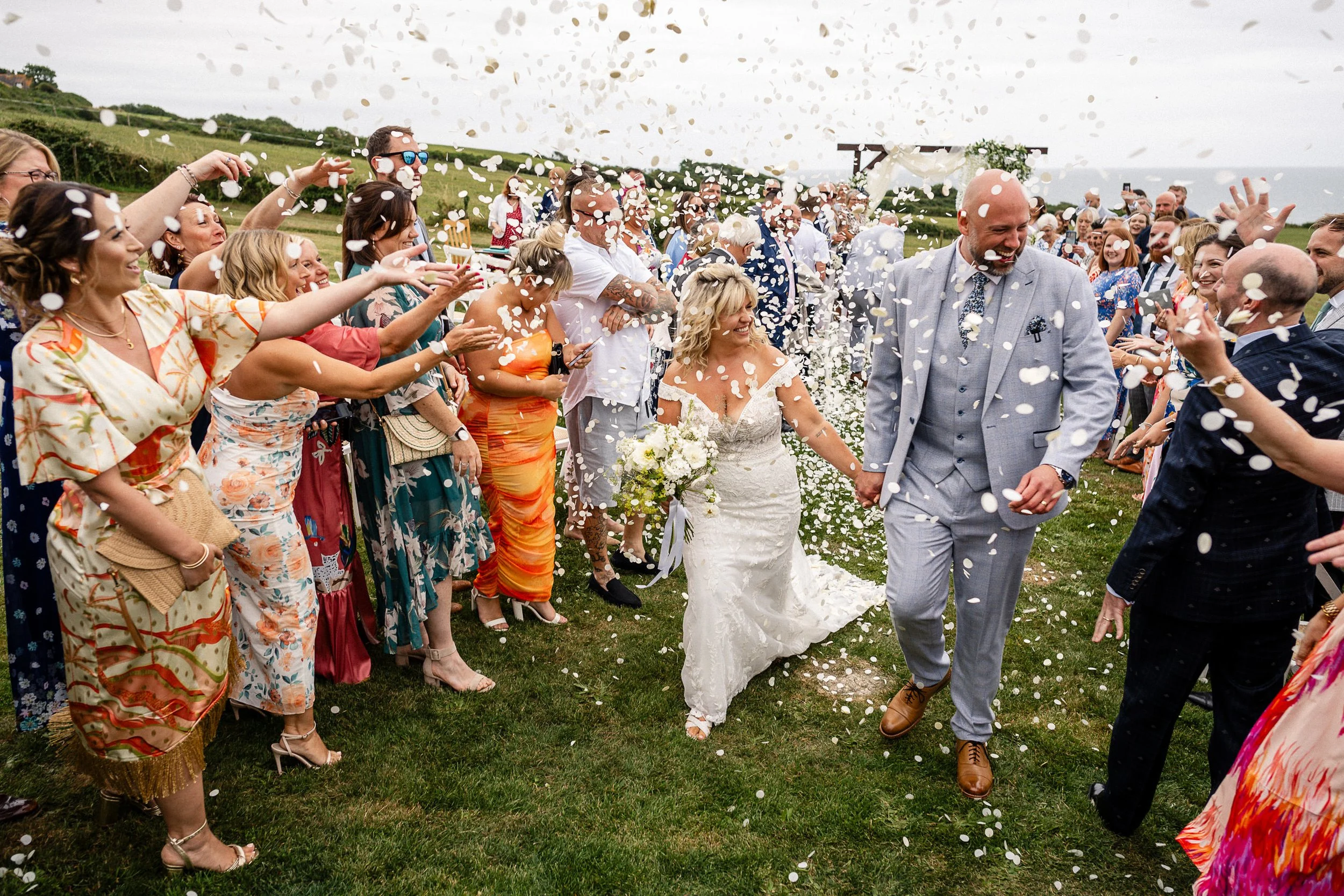 Newlywed bride and groom walk through cheering guests as white confetti fills the air at an outdoor wedding.