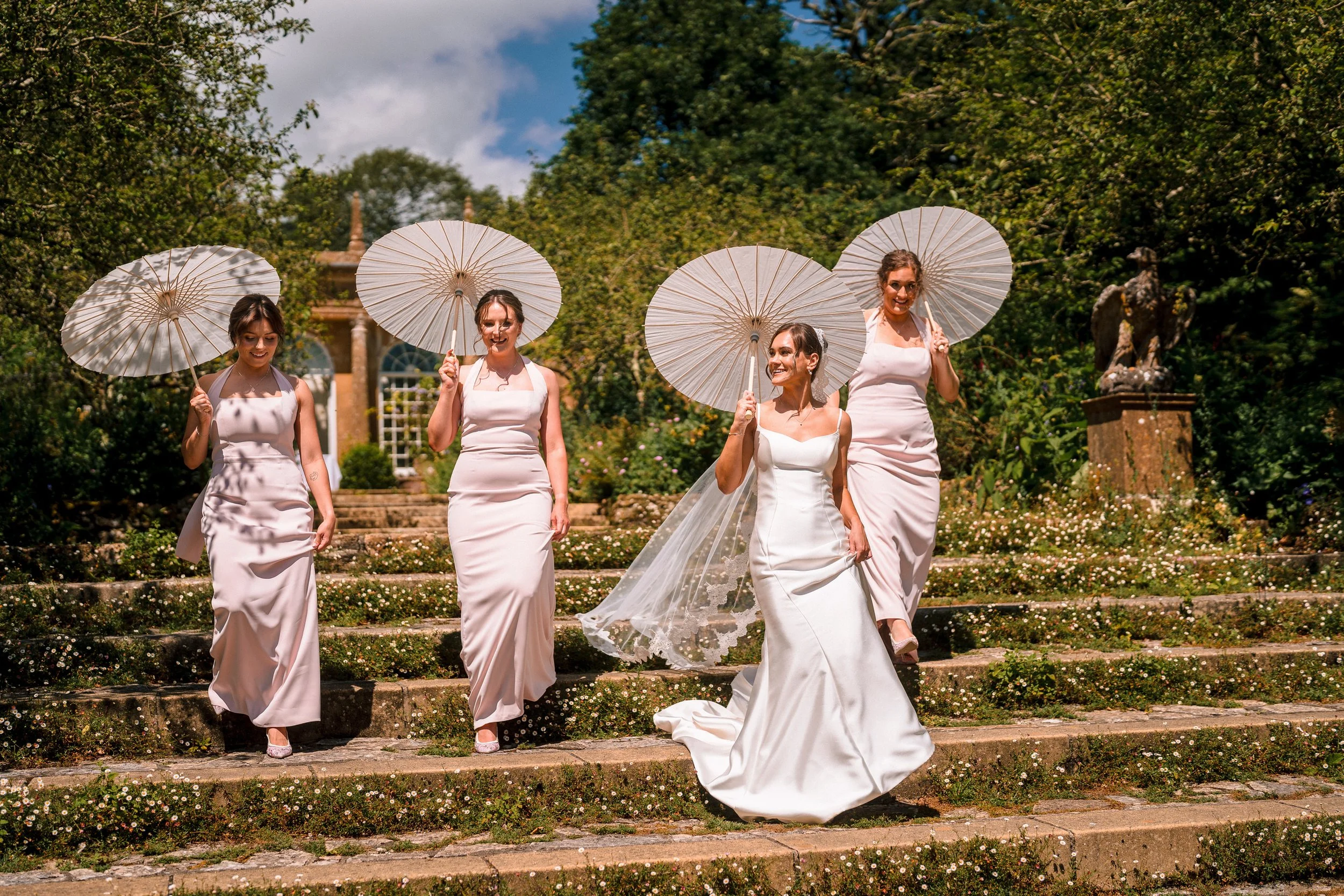 Bride and three bridesmaids in pale pink dresses carrying white parasols down garden steps at an outdoor wedding