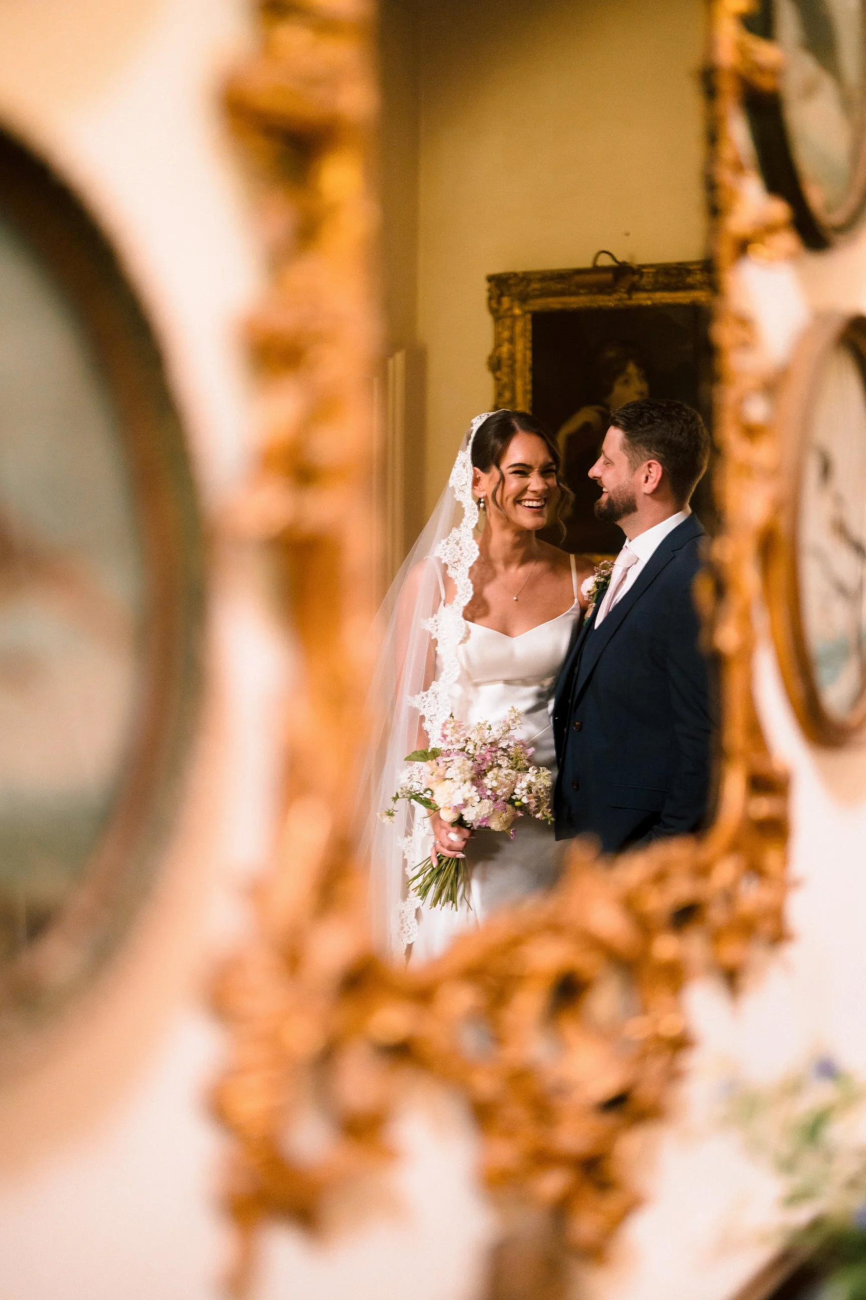 Smiling newlywed couple posing indoors, with the bride holding a pastel flower bouquet and blurred floral arrangements in the foreground.
