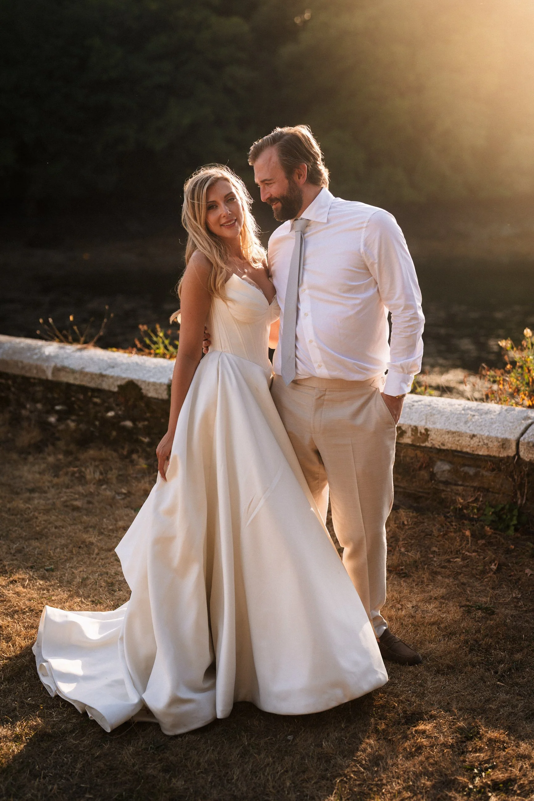 Newlywed couple posing outdoors at sunset, with the bride in a white wedding dress and groom in a white shirt and tie.