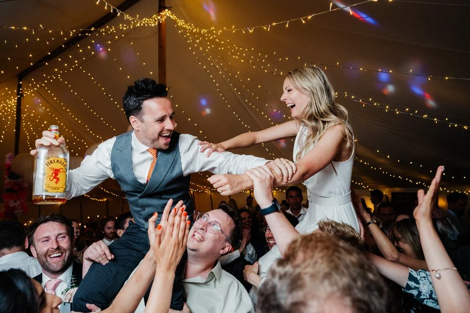 Wedding guests lift a laughing bride and groom during a lively reception under string lights, with the groom holding a Fireball whiskey bottle.