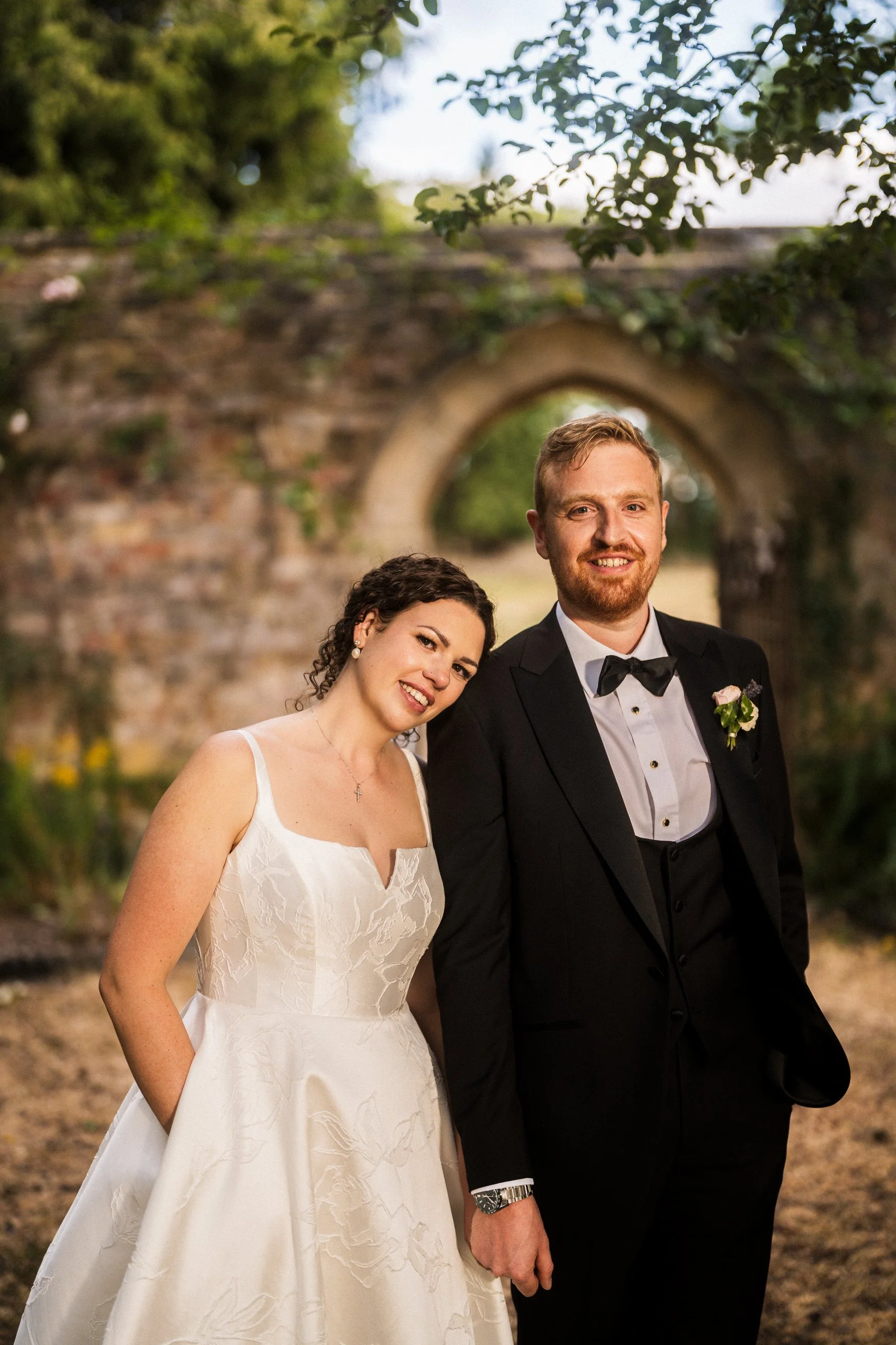 Newlywed couple posing outdoors beneath a leafy arch, bride in a white wedding dress and groom in a black tuxedo.