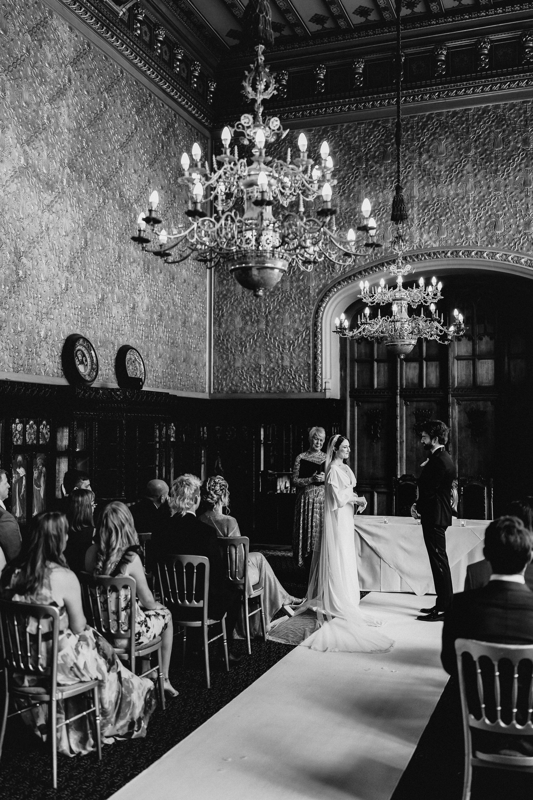 Black-and-white wedding ceremony in an ornate historic room with chandeliers, patterned walls, seated guests, and a bride and groom standing at the altar.