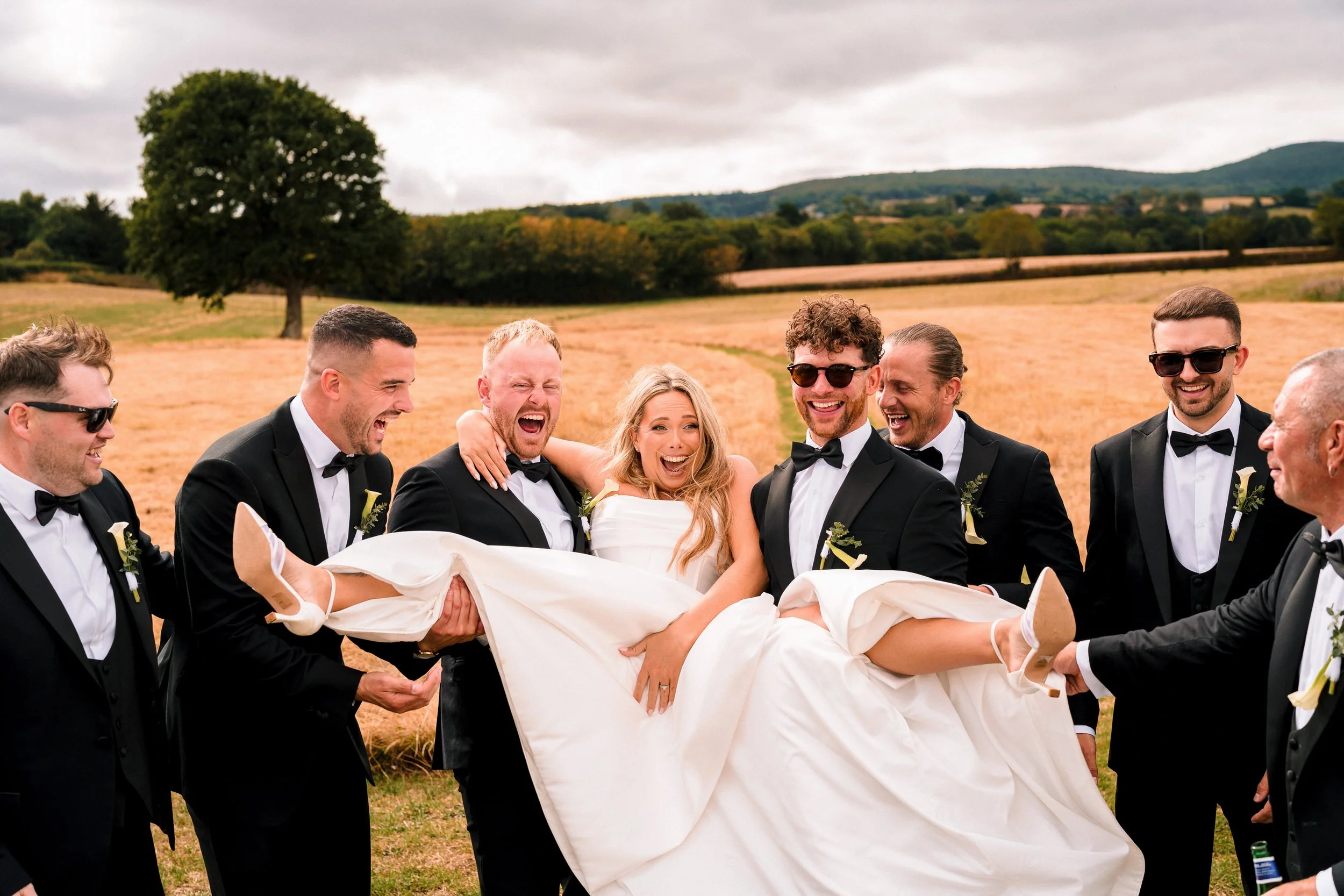 Bride in a white wedding dress being carried by laughing groomsmen in tuxedos across a countryside field