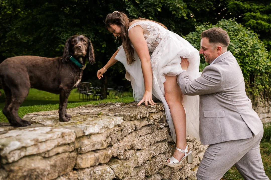 Groom helps bride climb a stone wall toward their dog during an outdoor wedding.