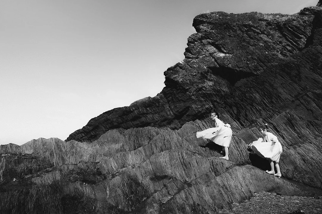 Black-and-white photograph of two women in flowing white dresses standing on rugged coastal rocks beneath a towering cliff.