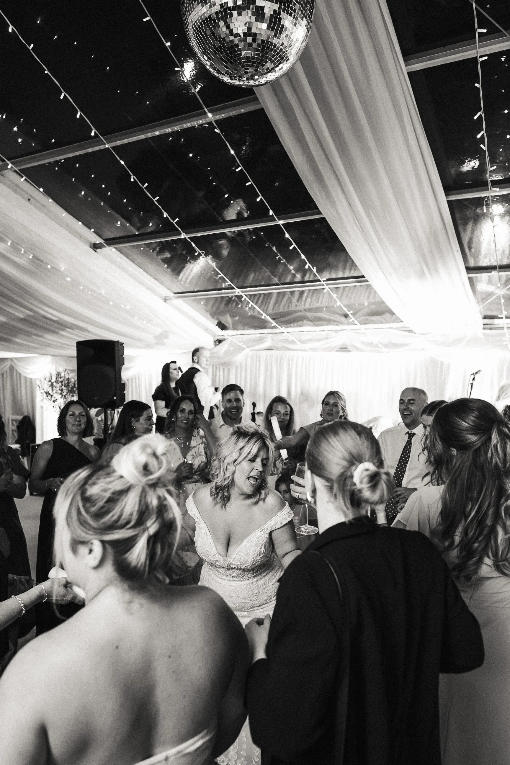 Bride dancing with wedding guests beneath draped fabric, string lights, and a disco ball at a lively reception