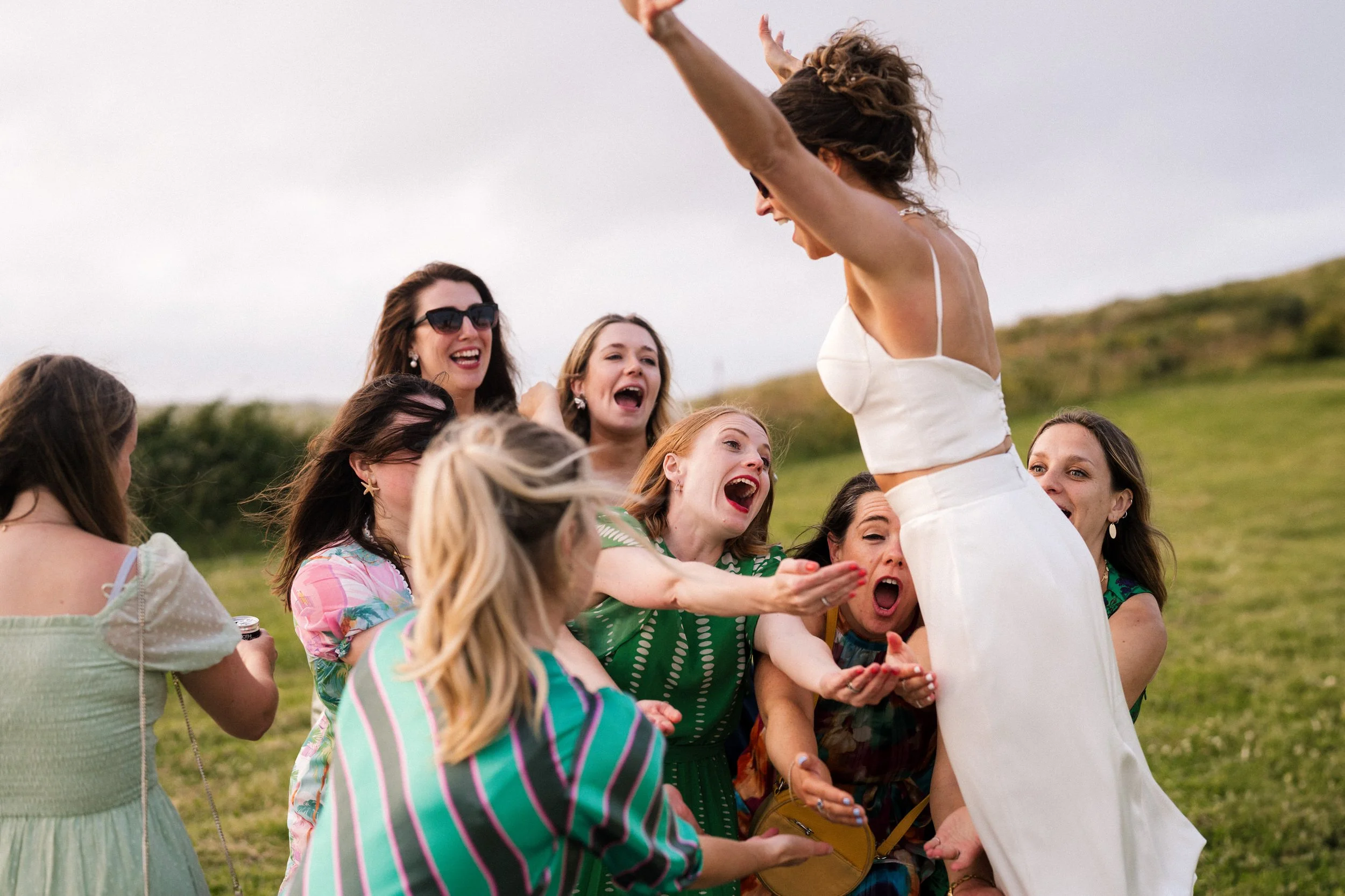 Group of women laughing and celebrating outdoors as they toss a woman in a white dress into the air.