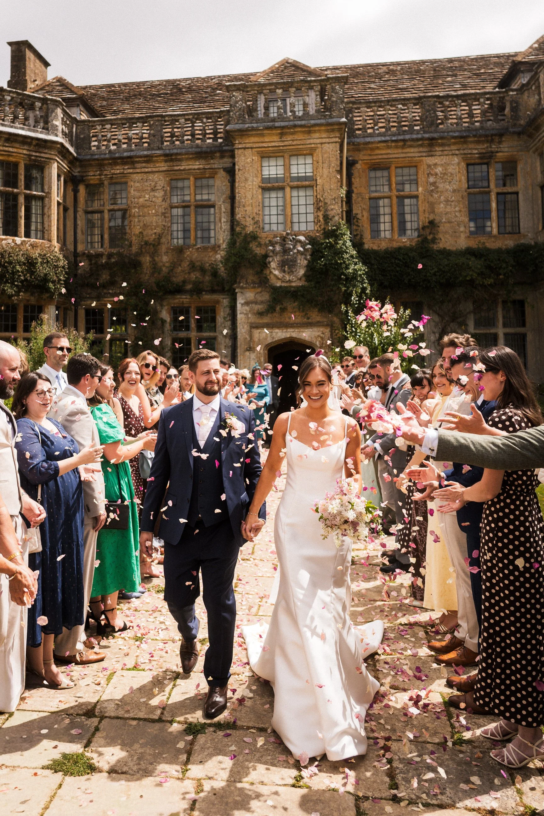 Newlywed couple walking through cheering guests as flower petals are thrown outside a historic stone country house