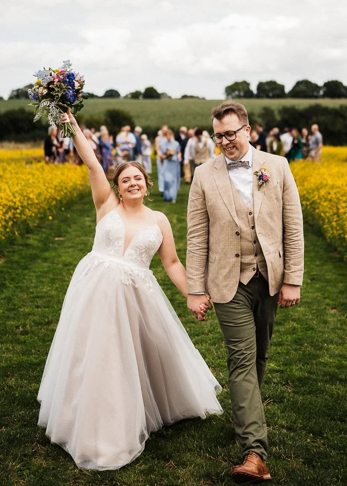 Newlywed couple walking hand in hand through a wildflower field, bride holding a bouquet and groom wearing a tan suit