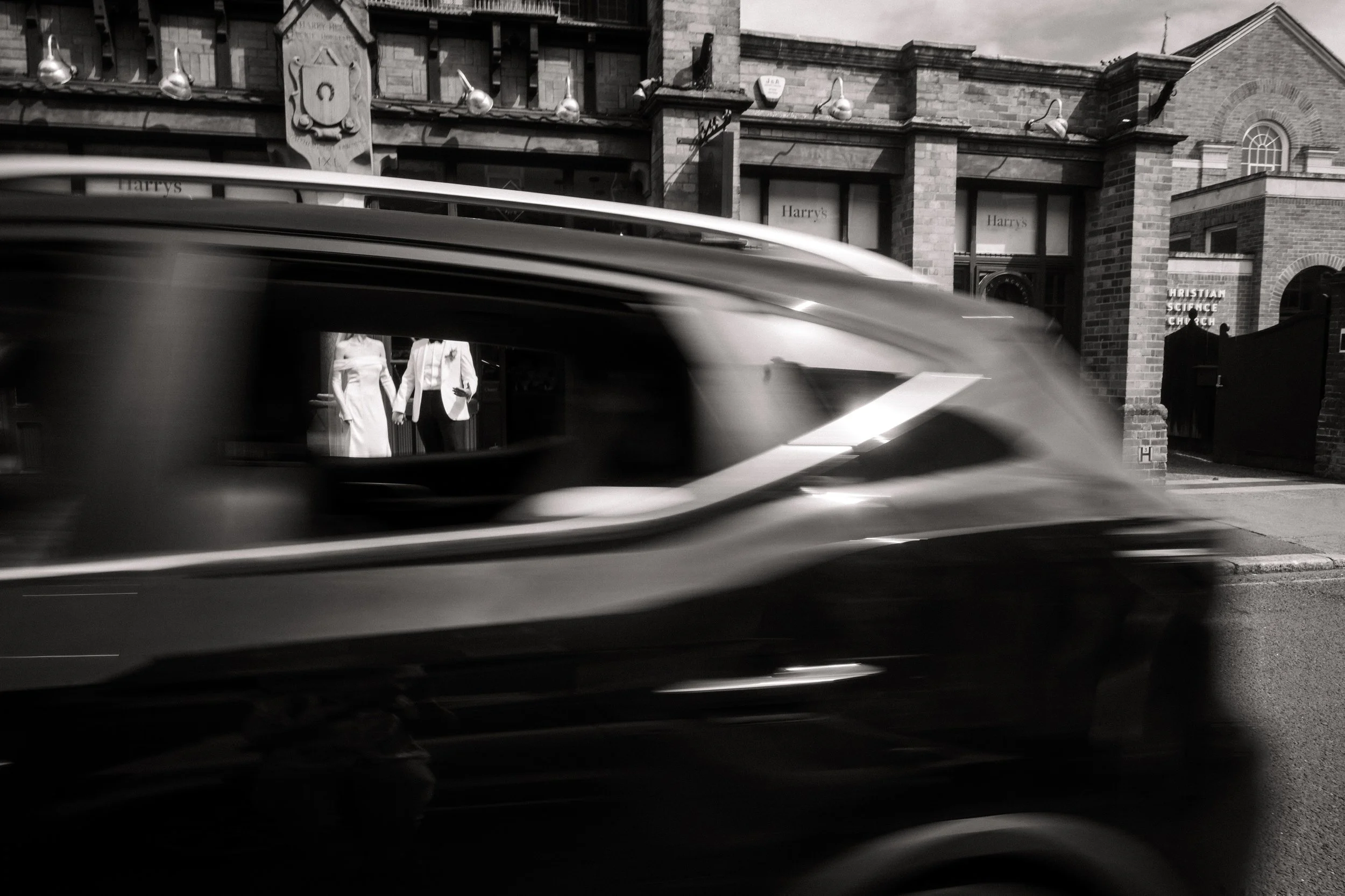 Black-and-white street scene with a blurred taxi passing Harry’s storefront and a Christian Science church.