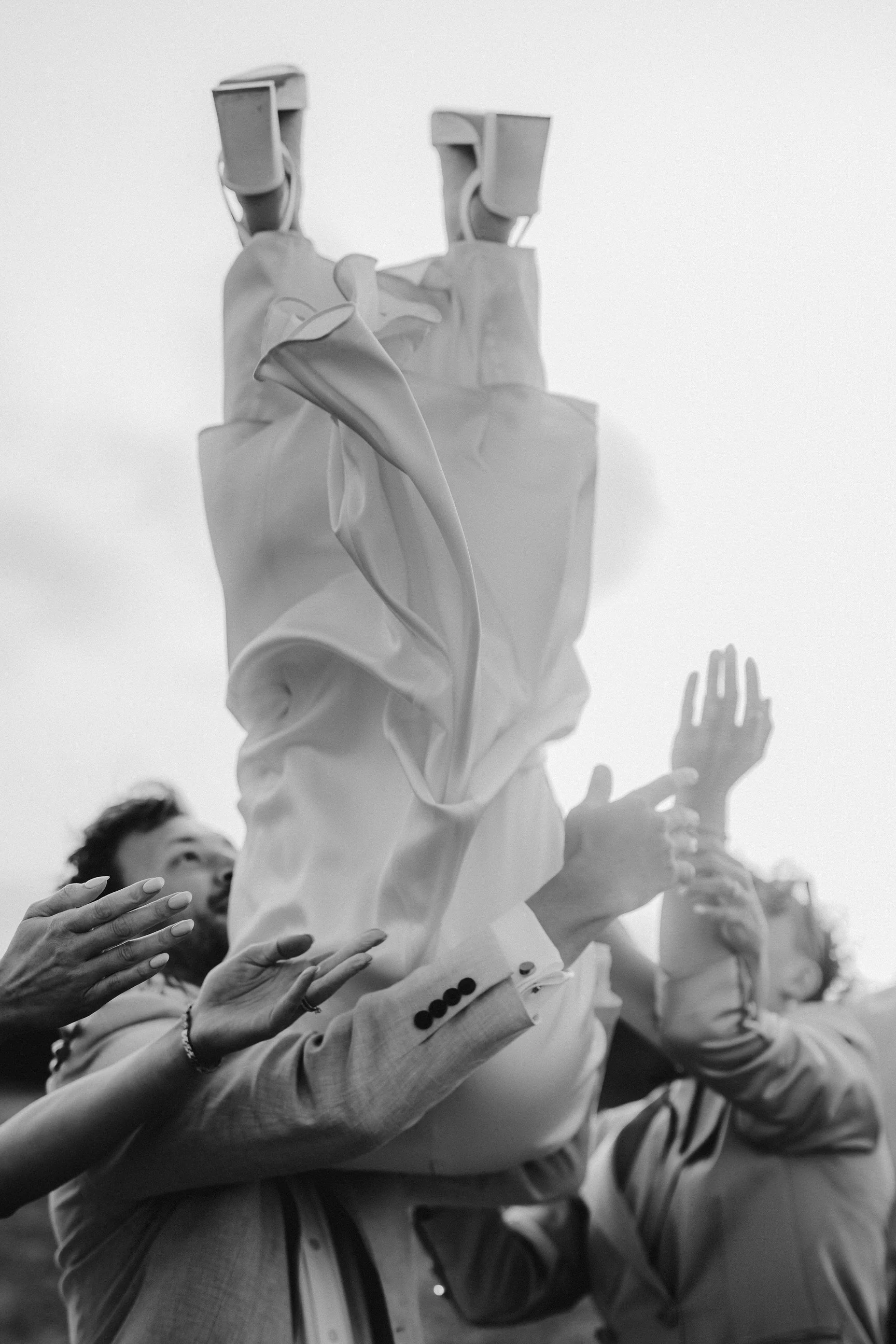 Black-and-white photo of a fashion model in a flowing white suit and sculptural hat, surrounded by photographers and raised hands.