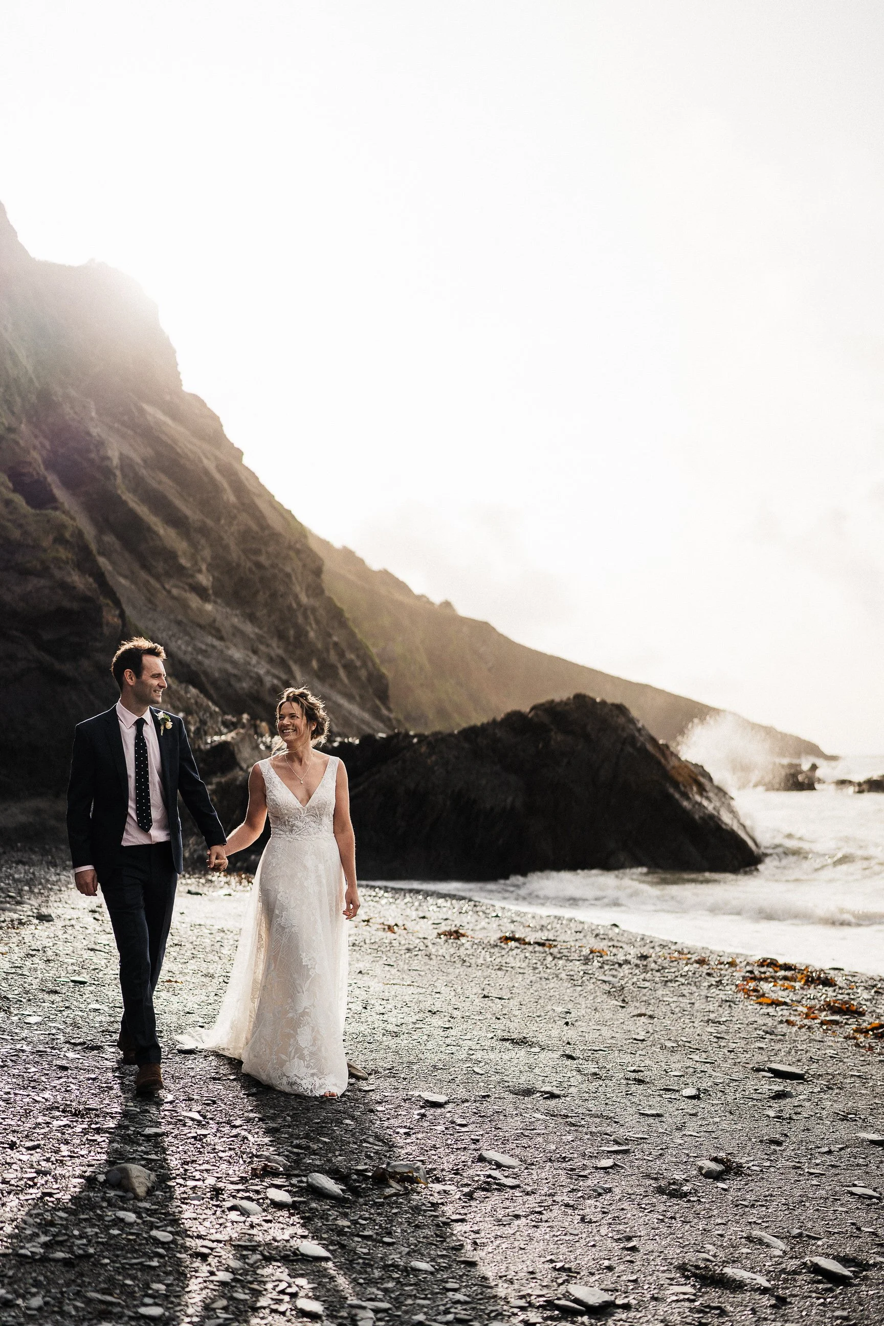 Newlywed couple walking hand in hand along a rocky beach beneath dramatic coastal mountains
