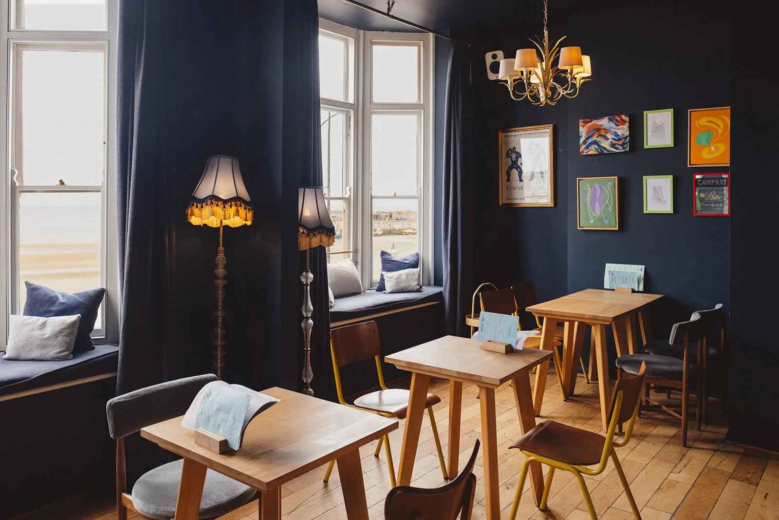 Cozy cafe interior with wooden tables and chairs, navy blue walls decorated with colorful artwork, large windows with dark curtains, window seats with pillows, and a chandelier with multiple lamps.